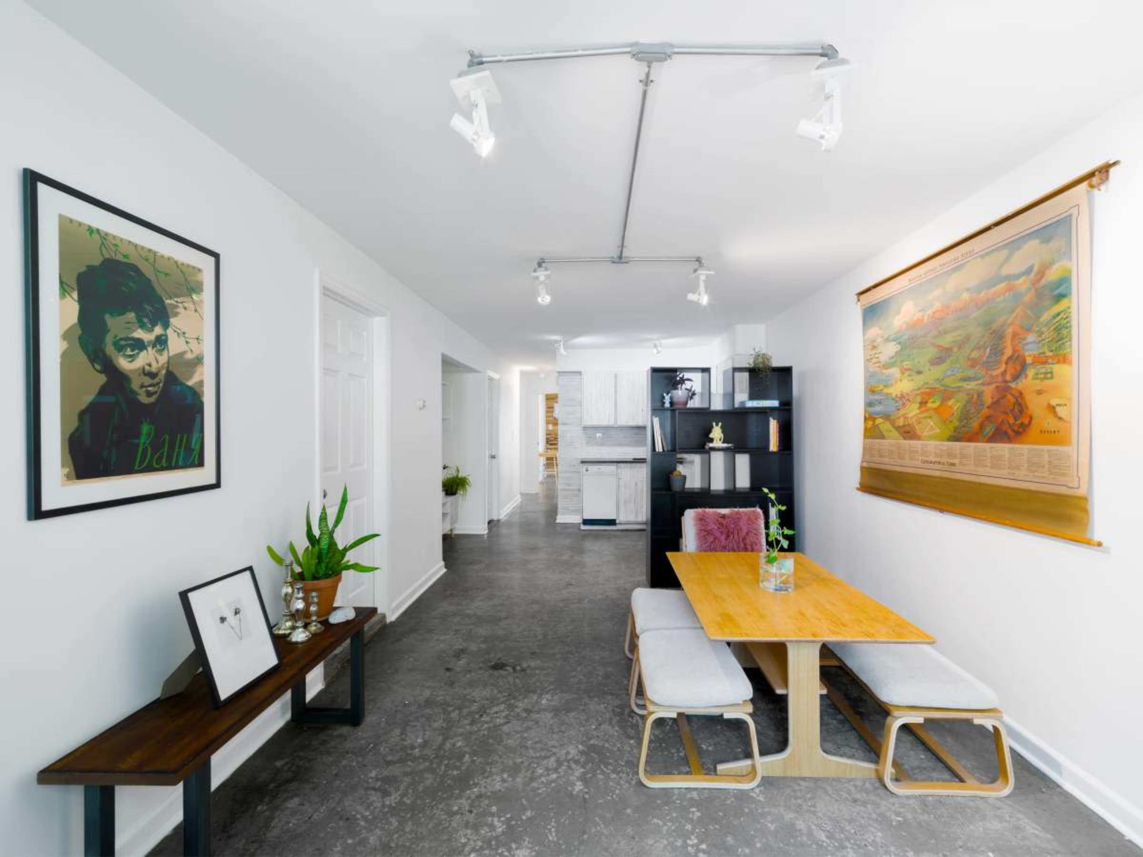 The image features a modern living space with a wooden dining table and chairs, a decorative wall with artwork, and open access to a kitchen area.