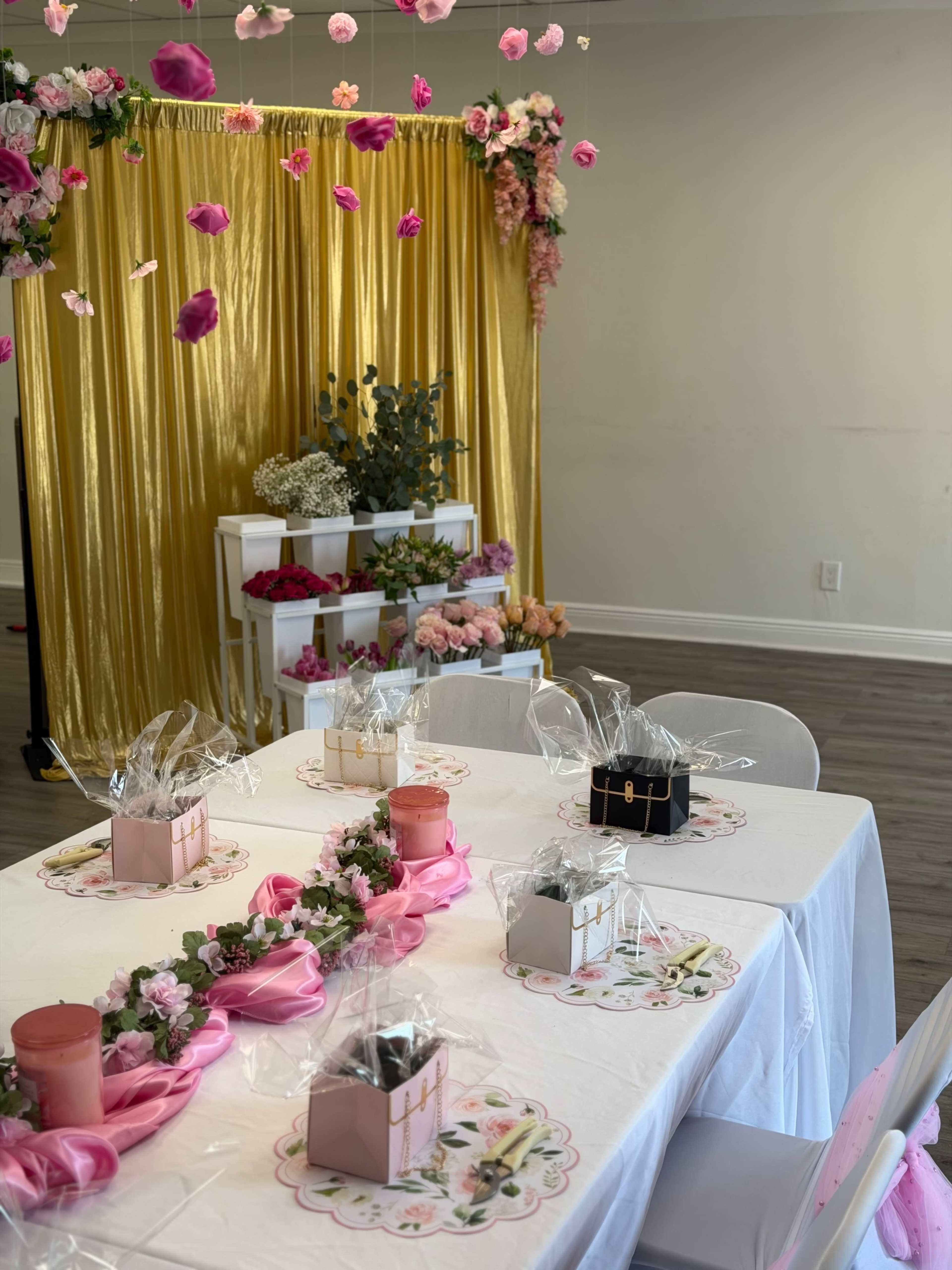 A decorated table is set with floral arrangements and gift boxes, surrounded by a backdrop of gold drapes and hanging flowers.