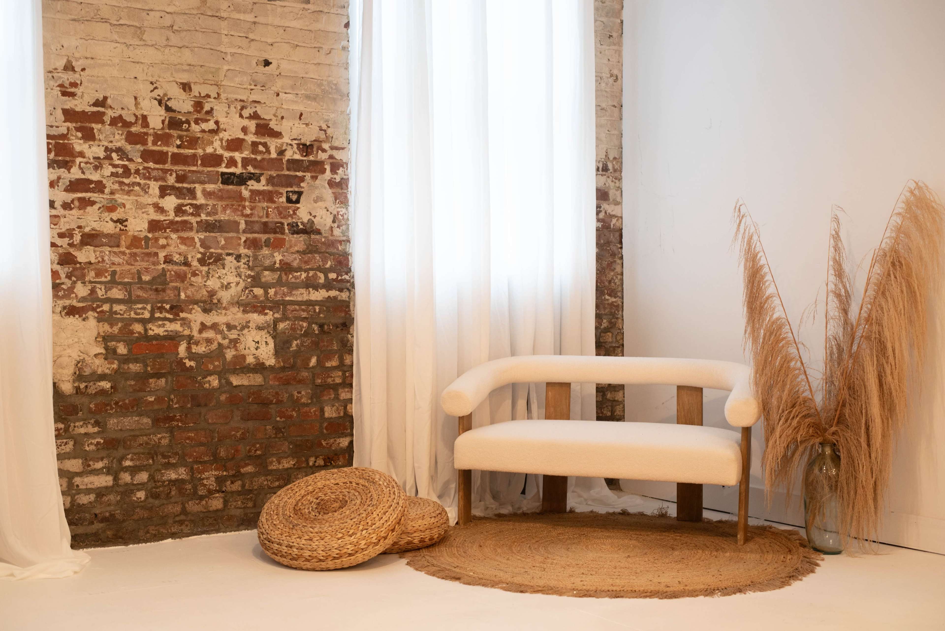 The scene features a minimalist room with a textured brick wall, a light-colored sofa, a round woven rug, and tall dried pampas grass in a vase.