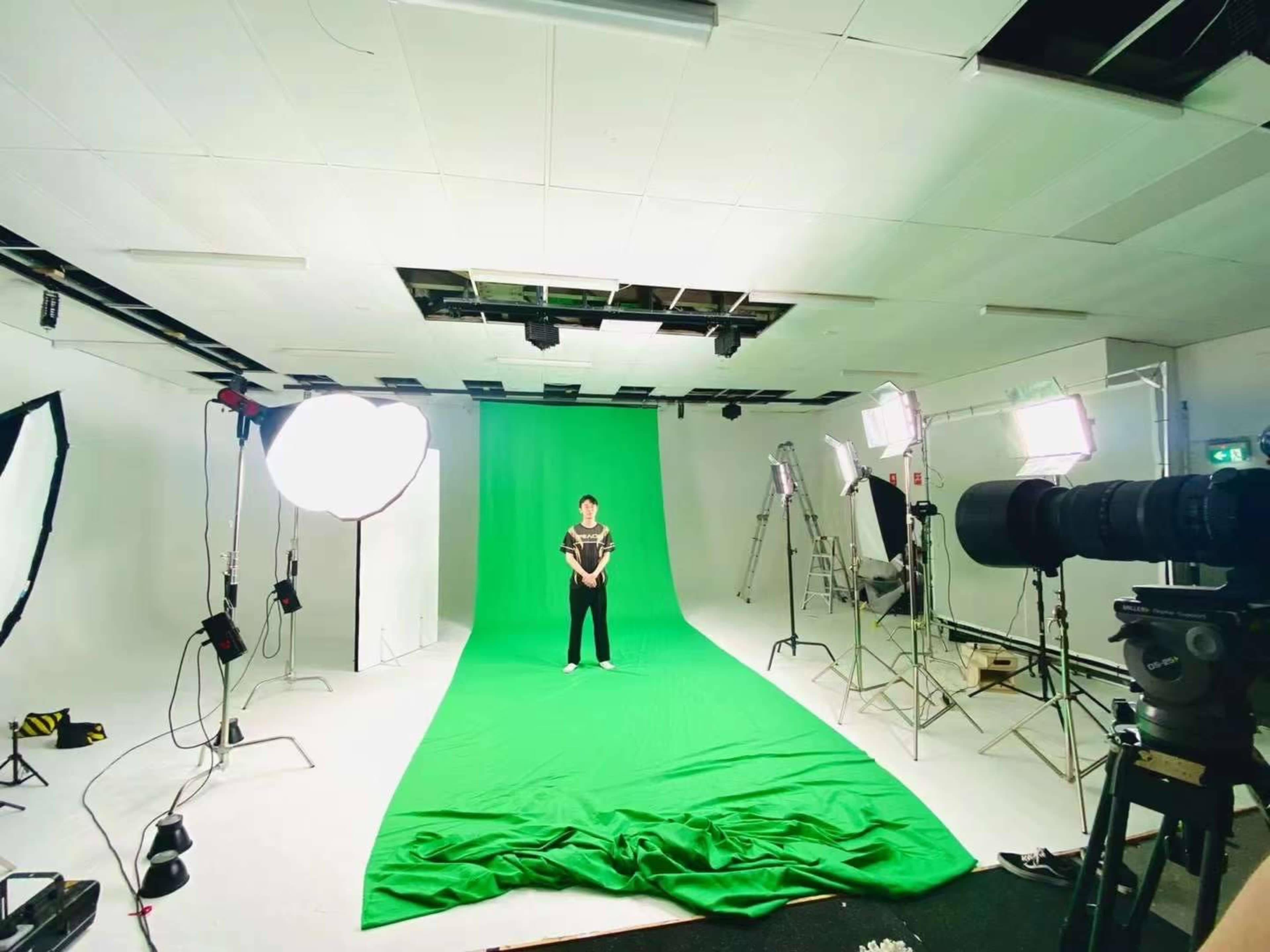 A green screen studio setup with a person standing in front of a large green backdrop, surrounded by multiple lighting equipment and cameras.