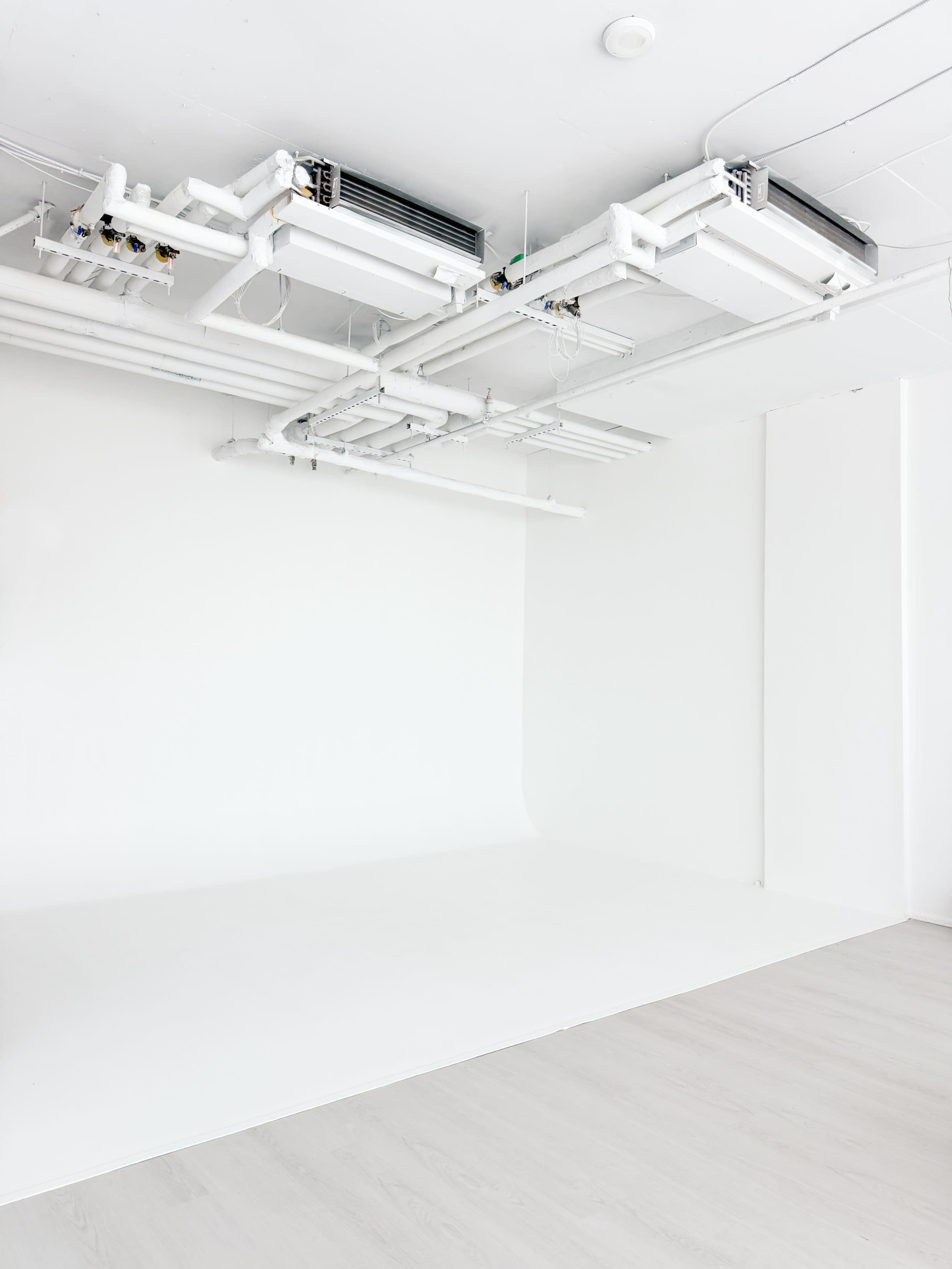 The image shows a minimalist room with a white wall, light wooden flooring, and exposed white pipes and air conditioning units on the ceiling.