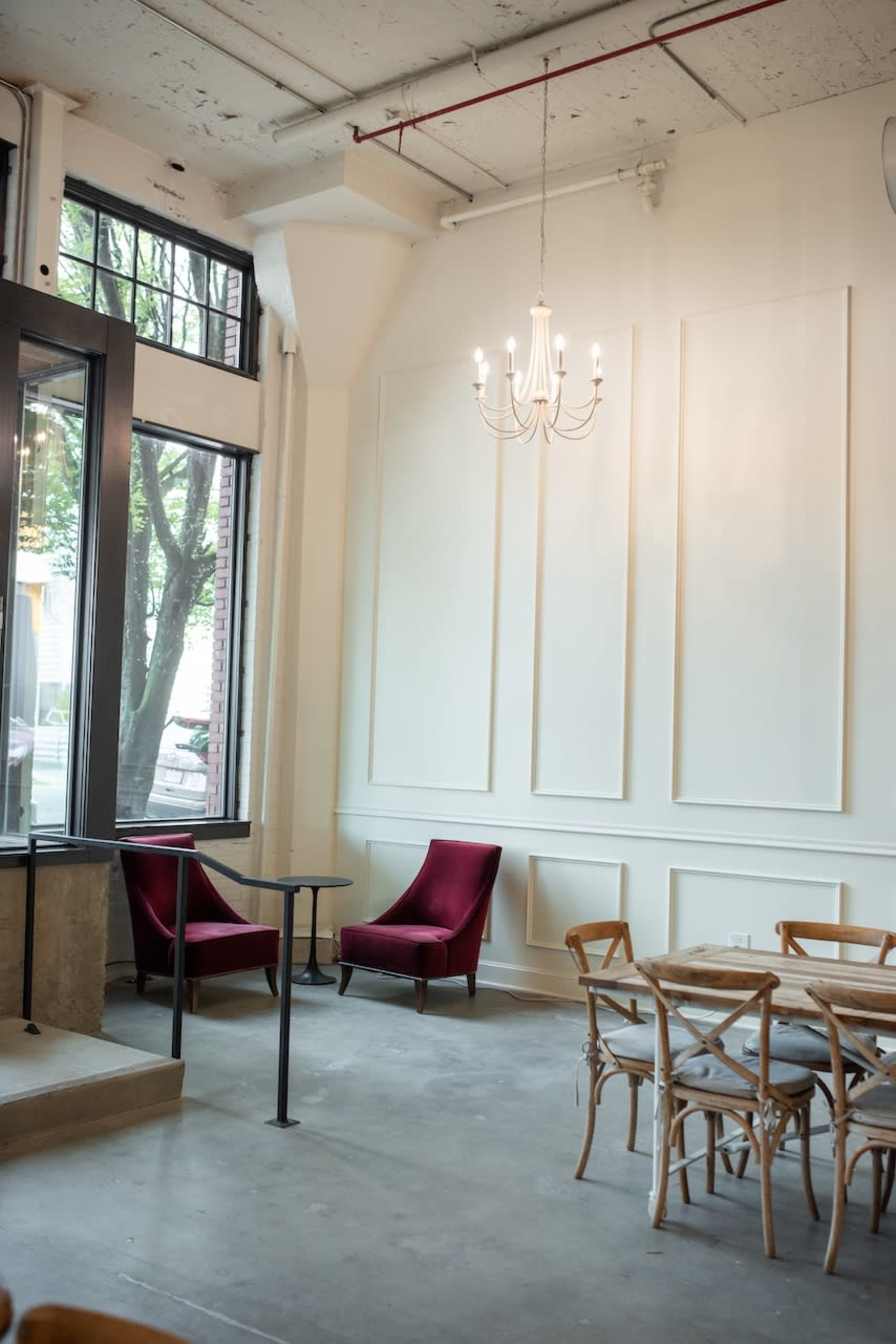 The image shows a modern café interior featuring two red armchairs near a small table, with a chandelier hanging from the ceiling and large windows allowing natural light to enter the space.