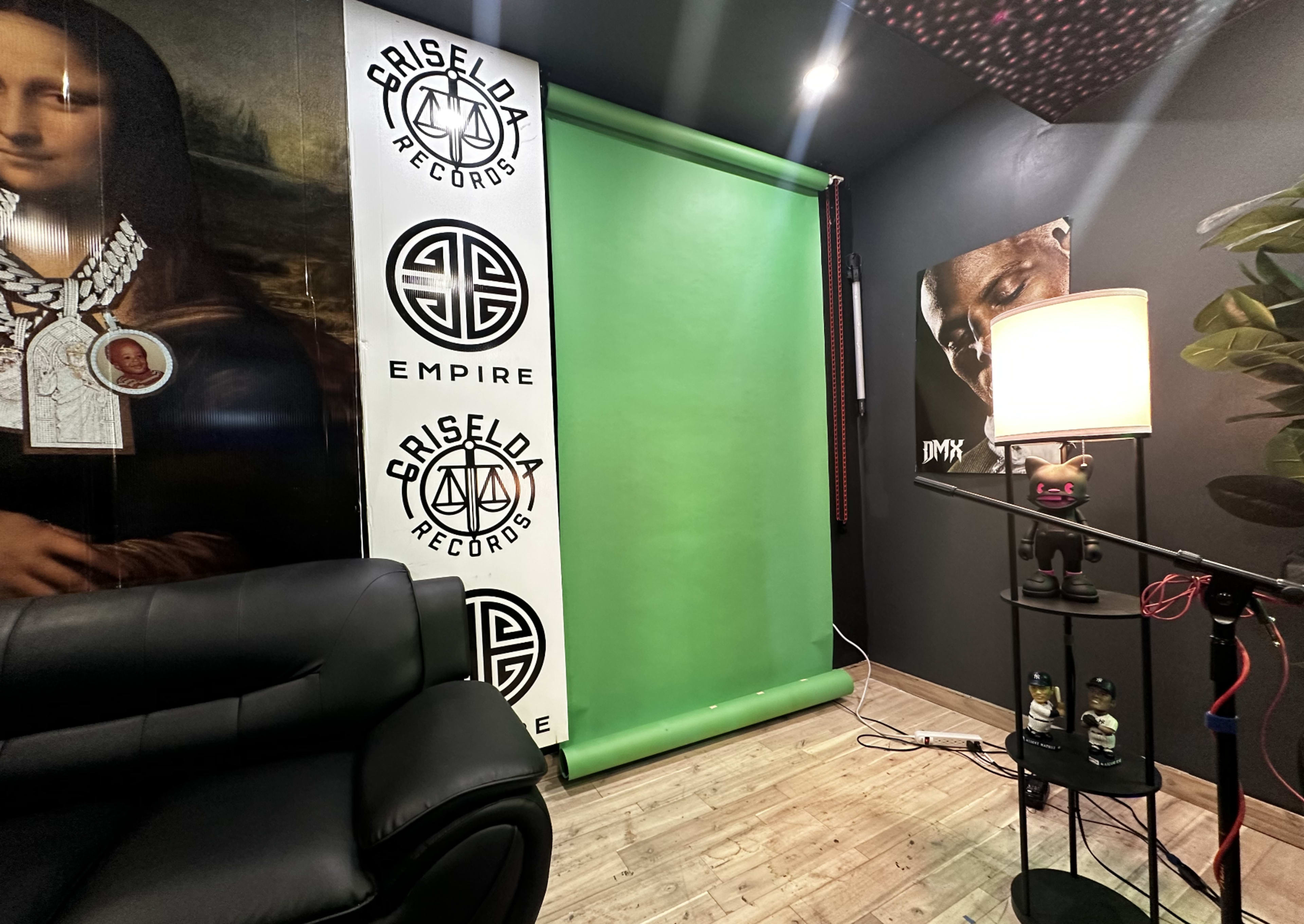 A corner of a room with a black leather couch, a green backdrop, brand logos on the walls, and a lamp beside a small table.