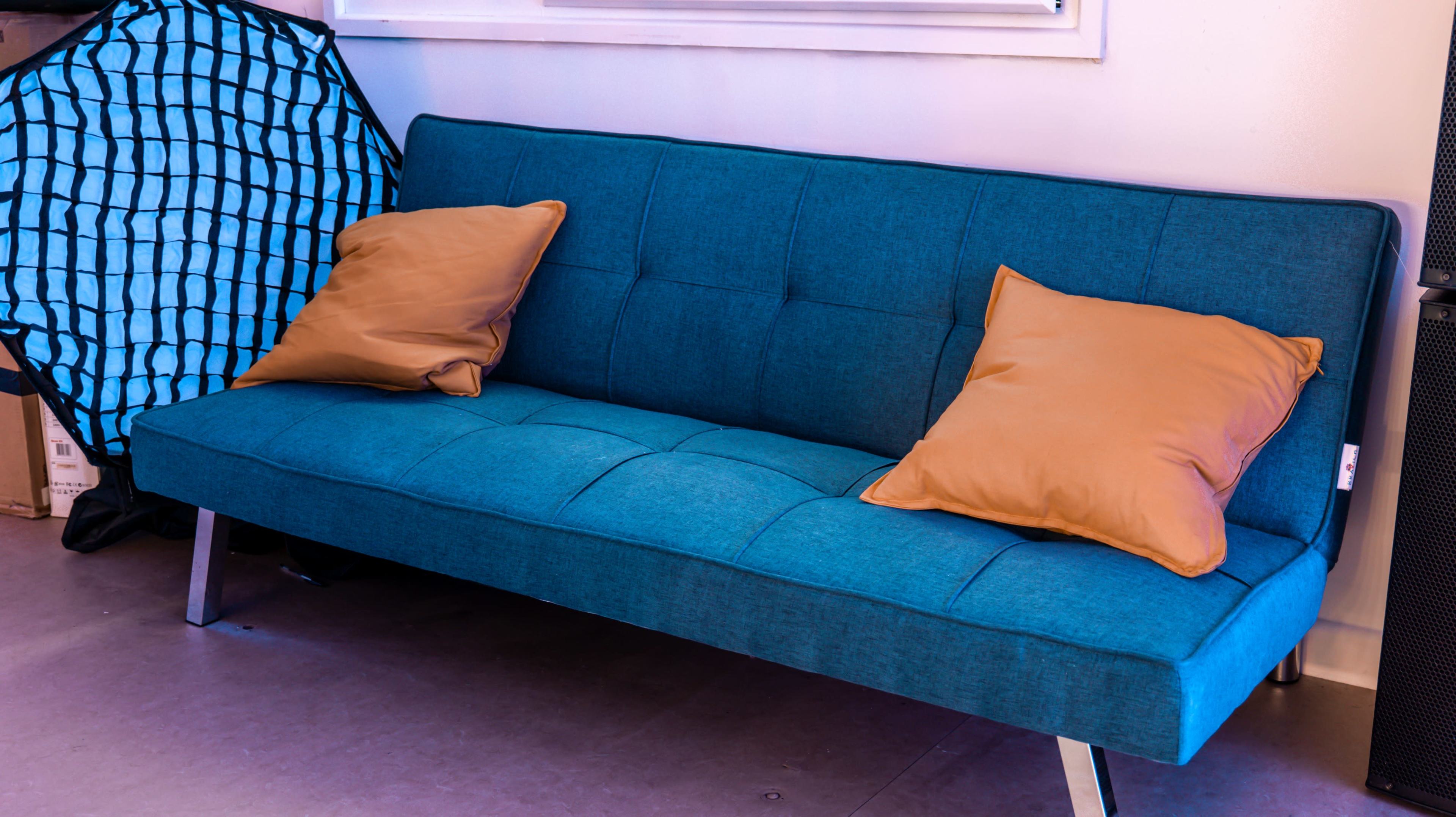 The image shows a blue couch with two orange pillows, set against a plain wall in a room with a large grid-patterned object in the background.