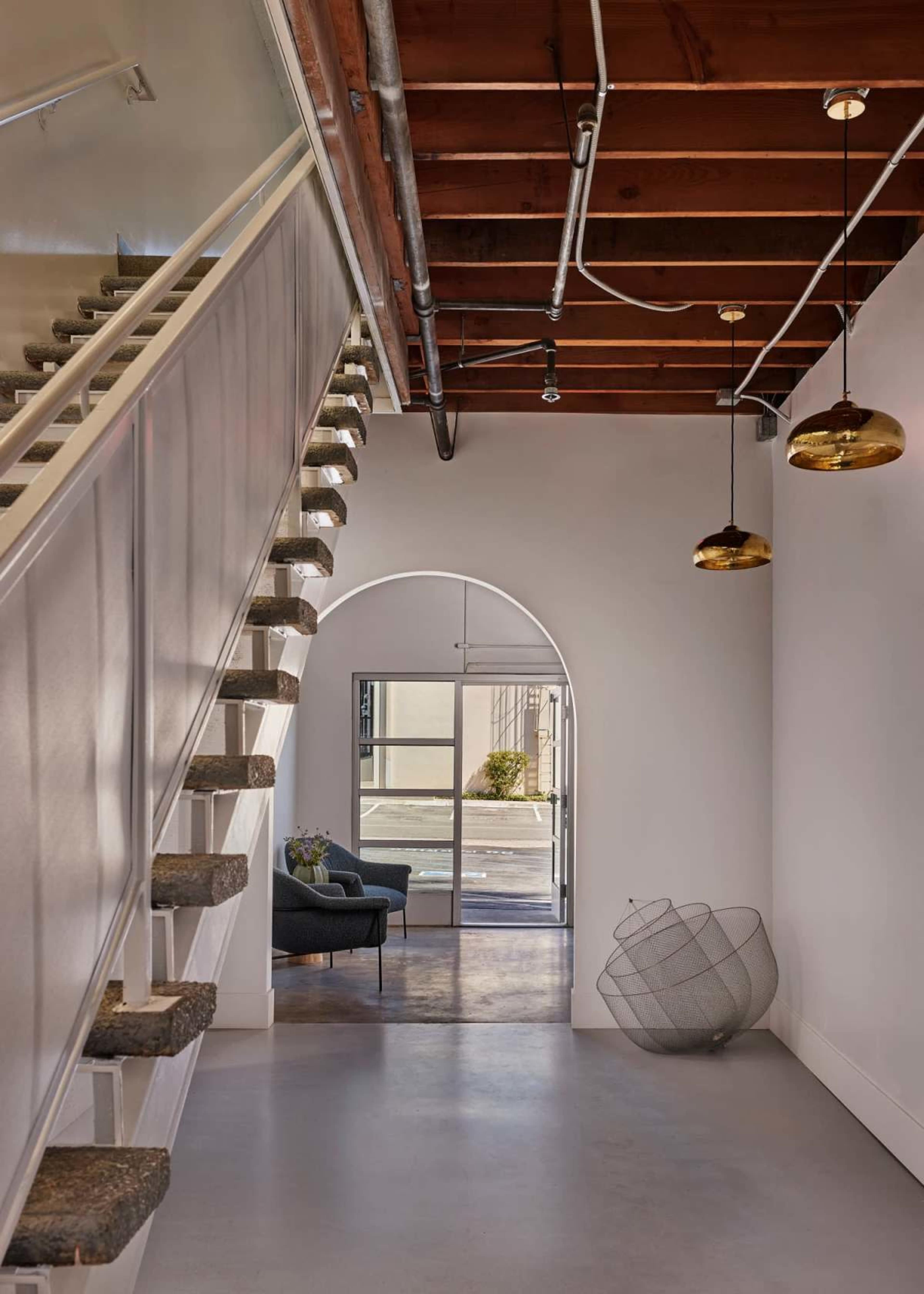 A staircase with a wooden railing leads up to a hallway that features a large archway and modern decor, including a pair of pendant lights and a wire basket.