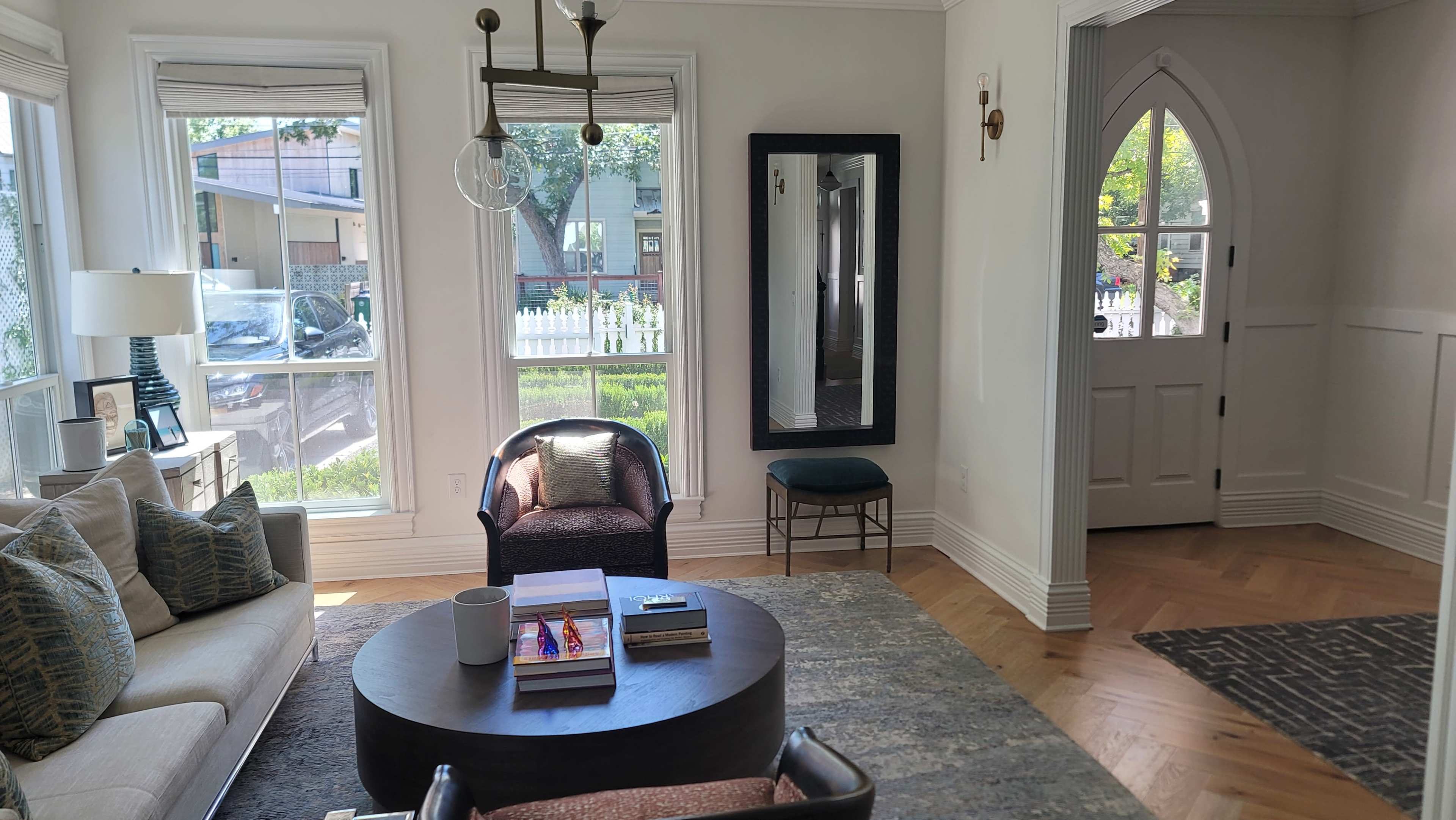 A living room features a round coffee table with books, two armchairs, and a large mirror, while an arched entryway leads outside.