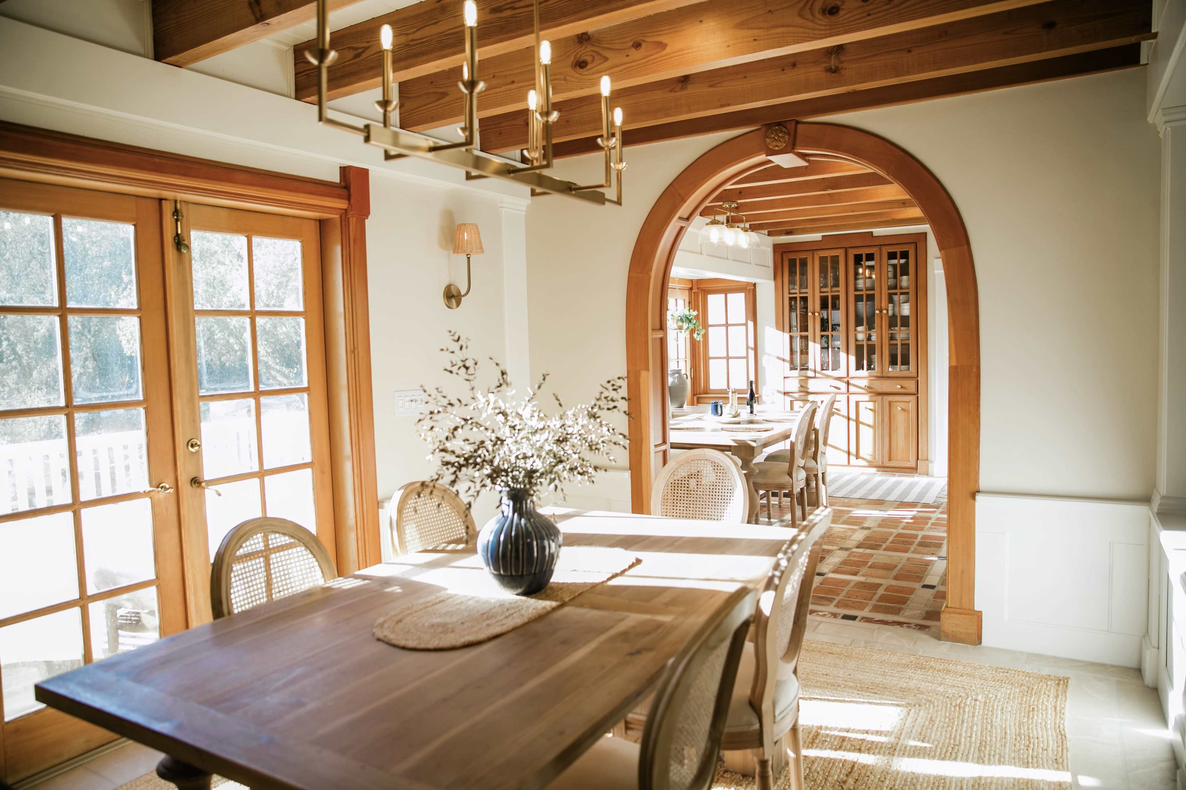 A sunlit dining area features a wooden table with a vase of flowers, surrounded by chairs, leading to a spacious kitchen area.