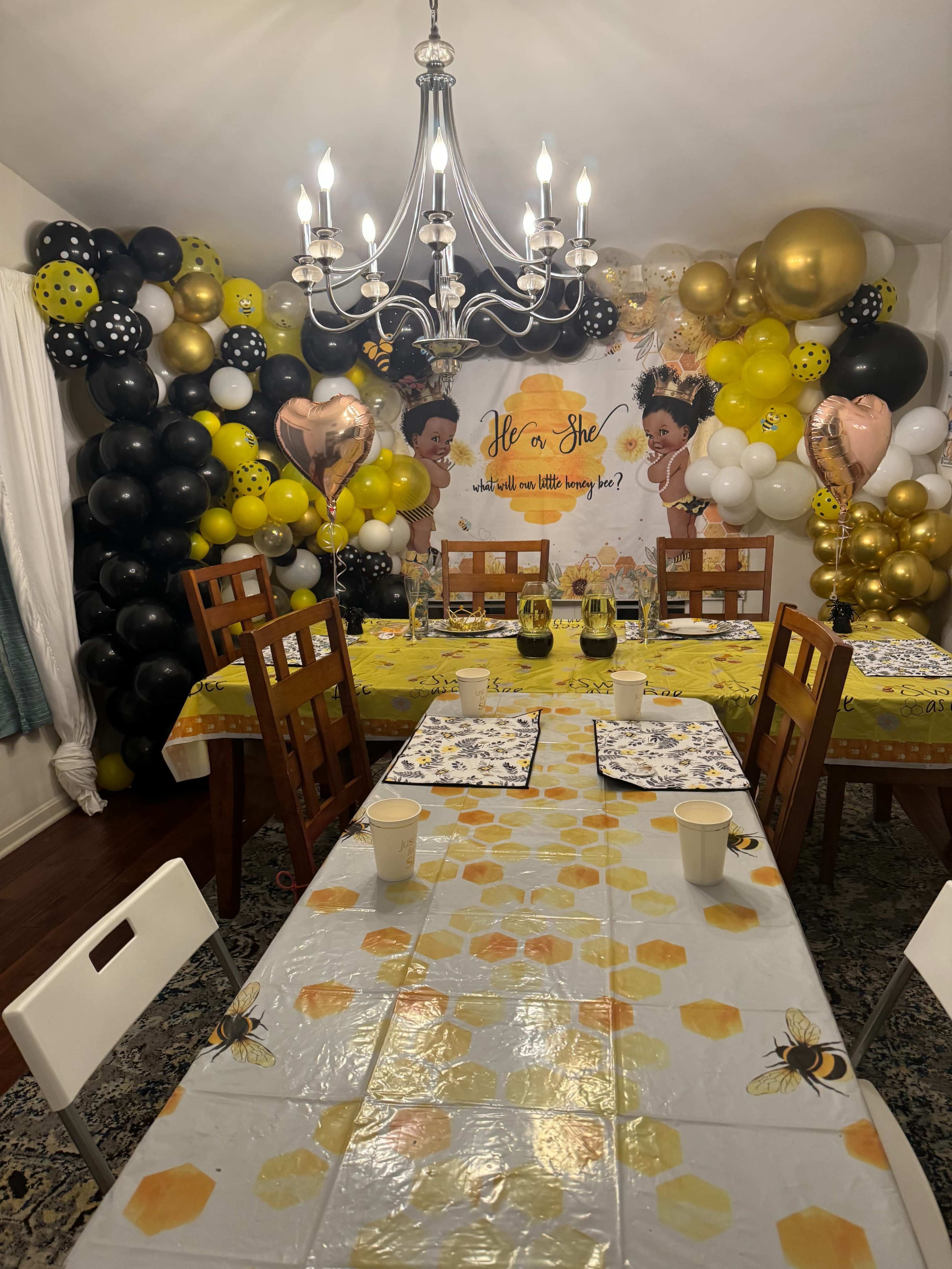 A long dining table is set with yellow and bee-themed decorations, surrounded by a balloon arch and a backdrop featuring a celebratory theme.