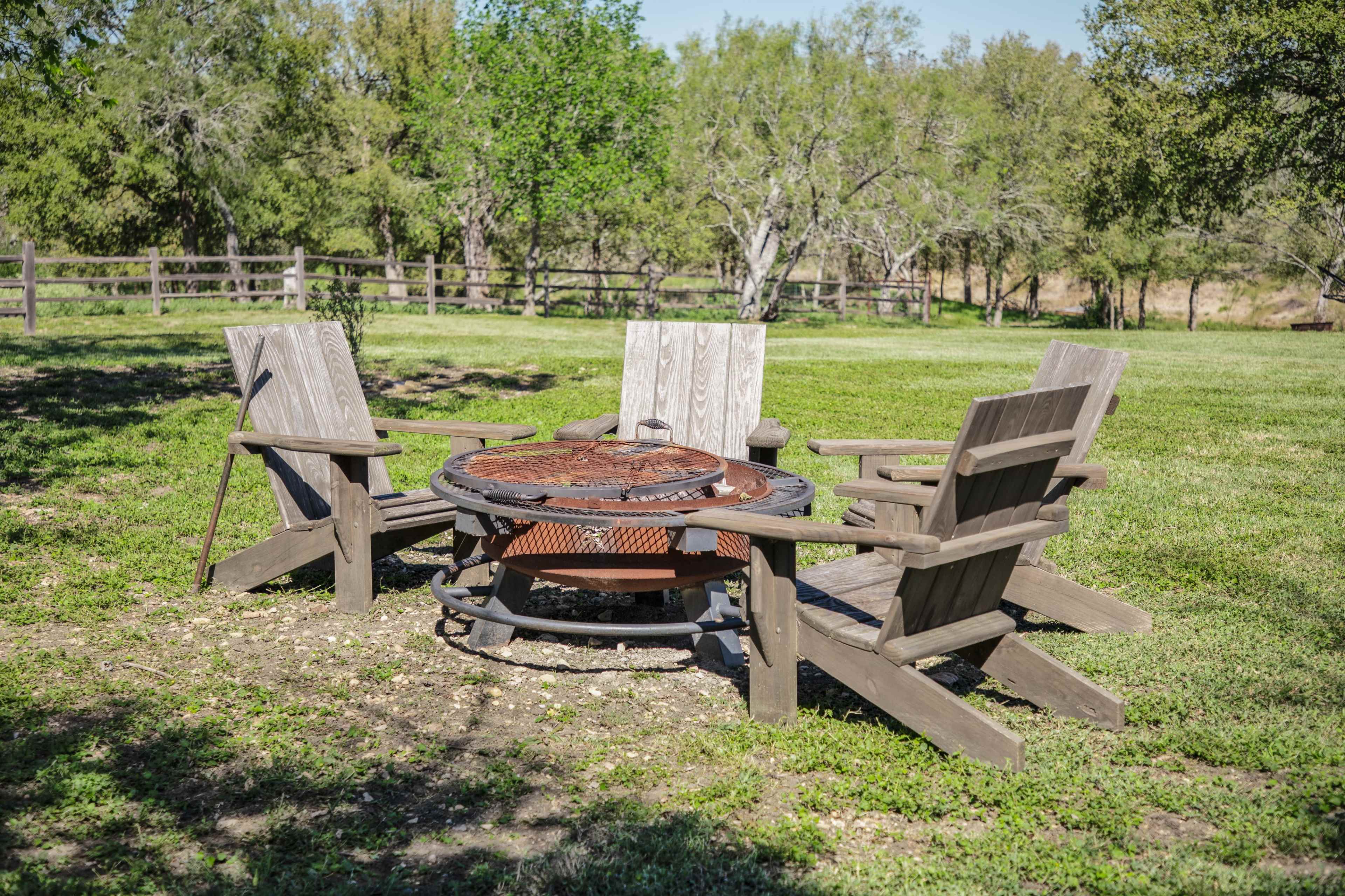 A circular fire pit surrounded by four wooden chairs sits in a grassy area with trees and a fence in the background.