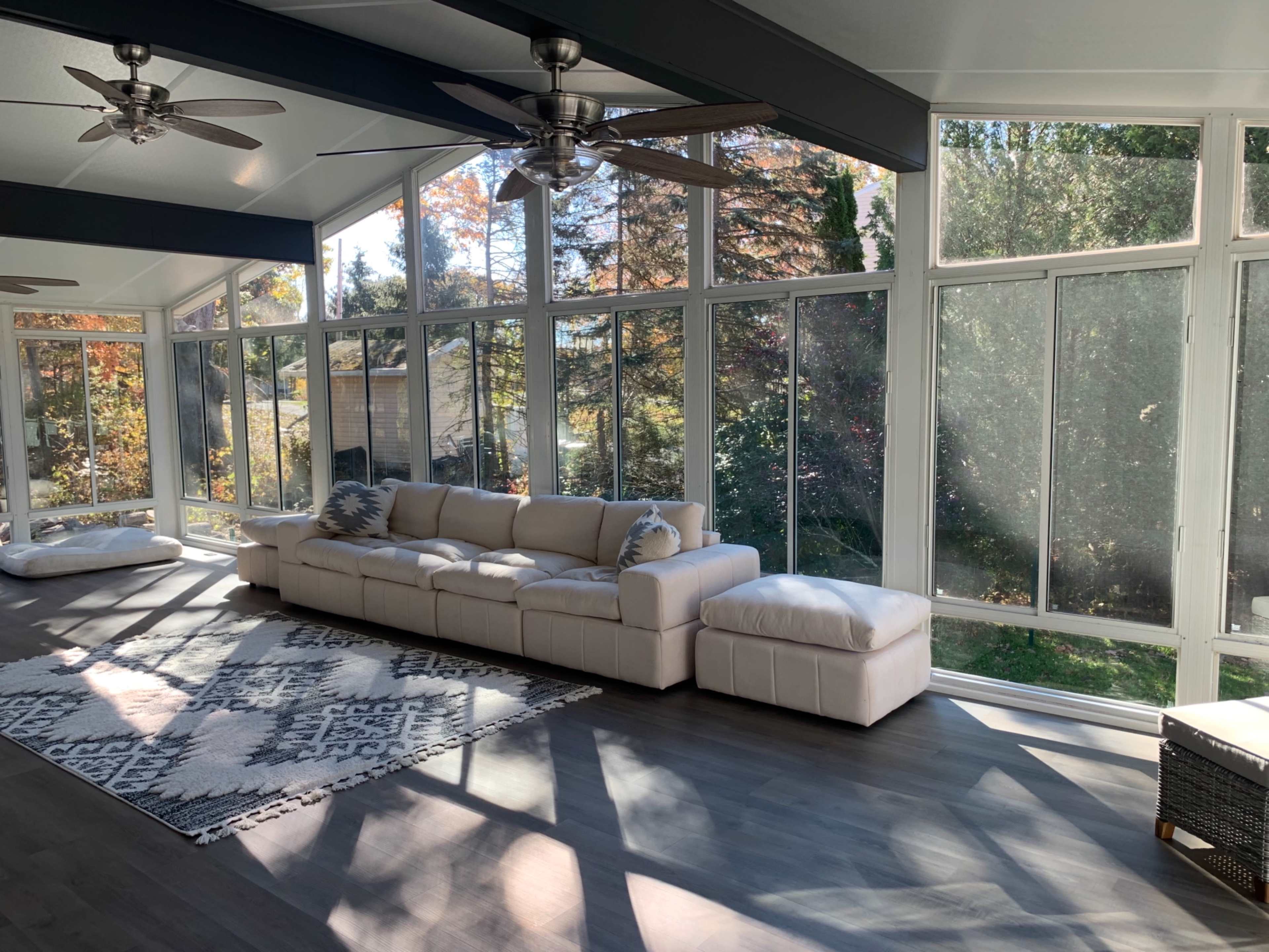 A sunlit sunroom features a large sectional sofa, ceiling fans, and a patterned rug against a backdrop of large windows.