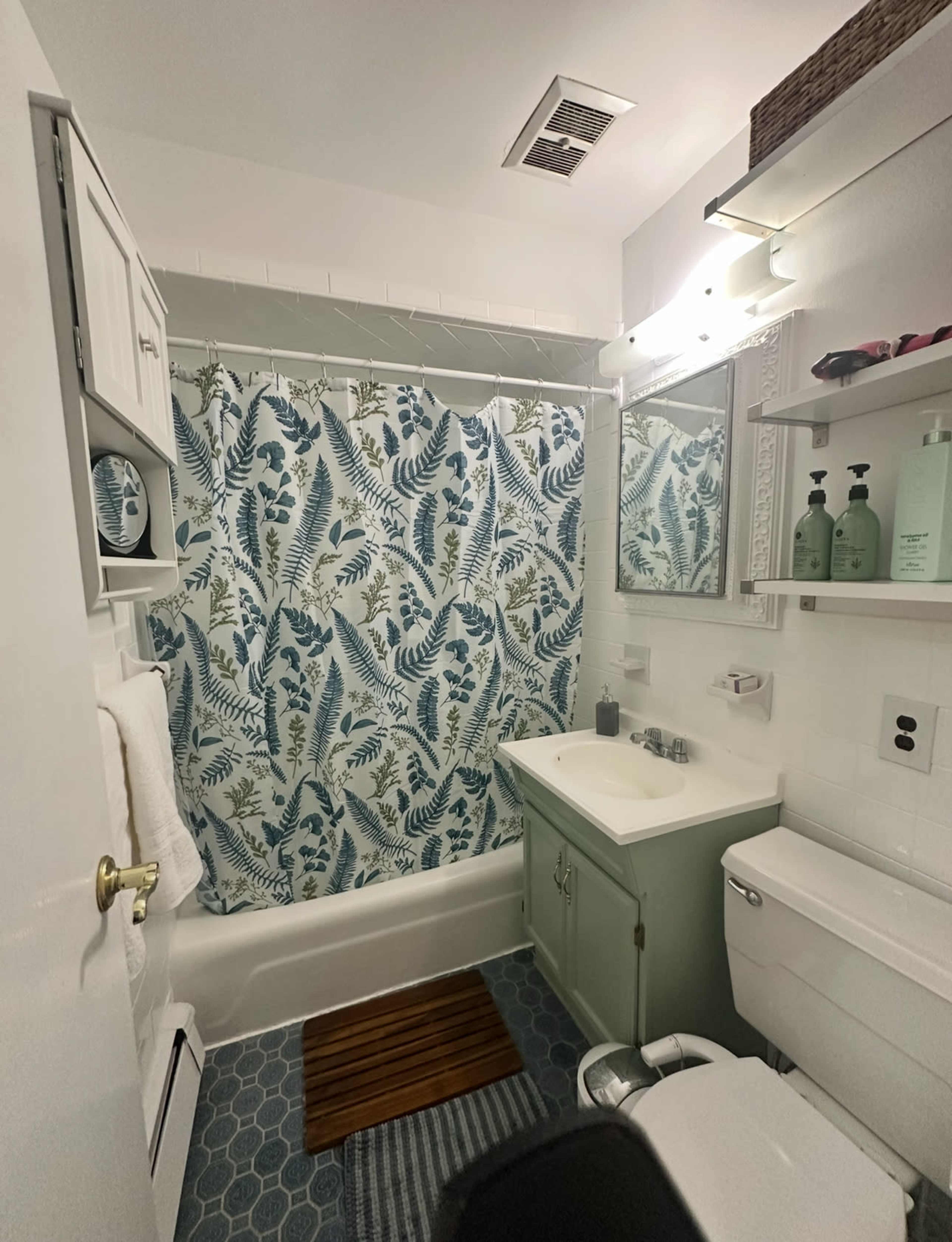 The image shows a small bathroom featuring a bathtub with a floral shower curtain, a light green vanity, and shelves with toiletries.