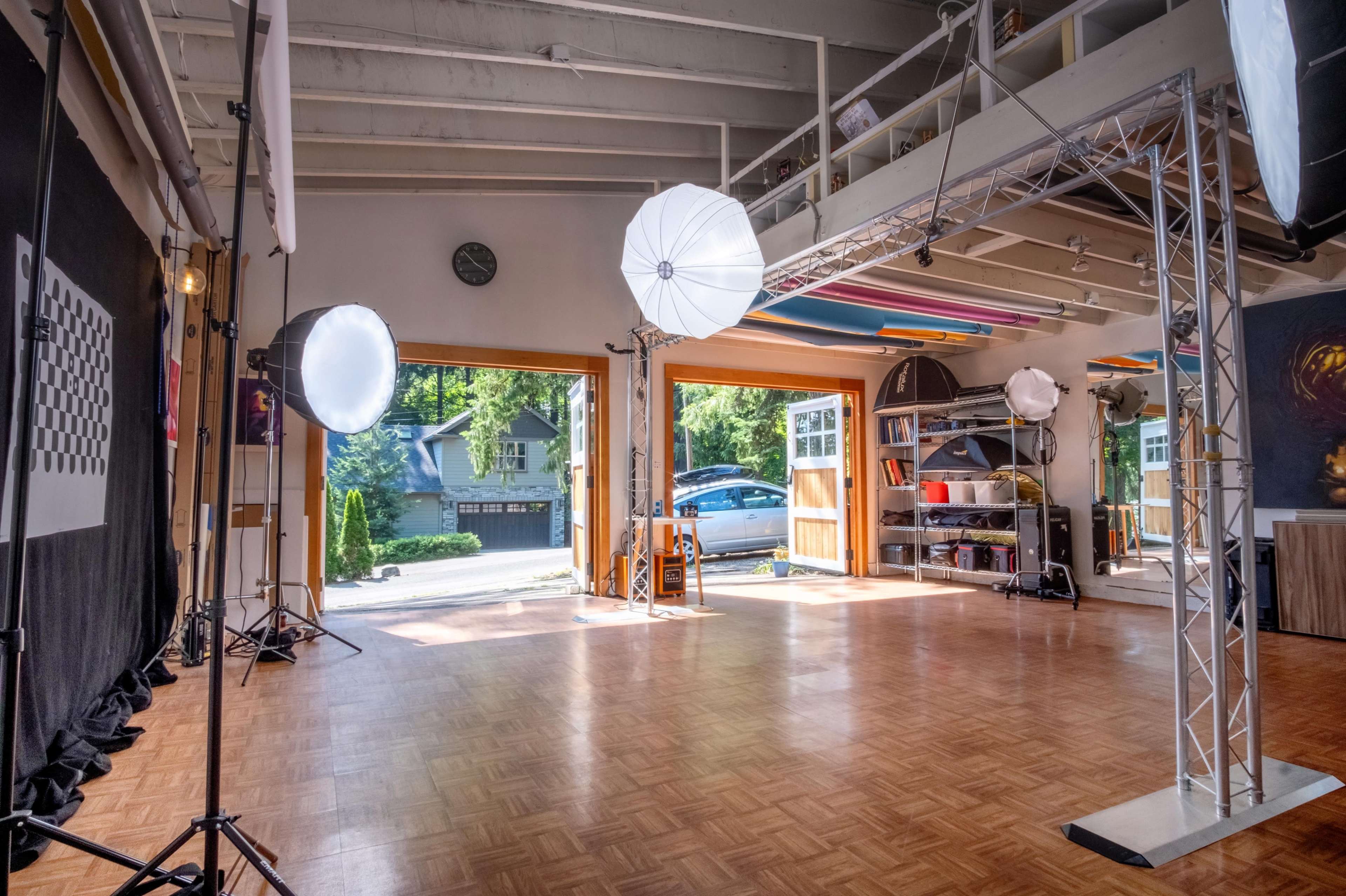 A photography studio features large windows, lighting equipment, and a wooden floor, with a view of a house outside.