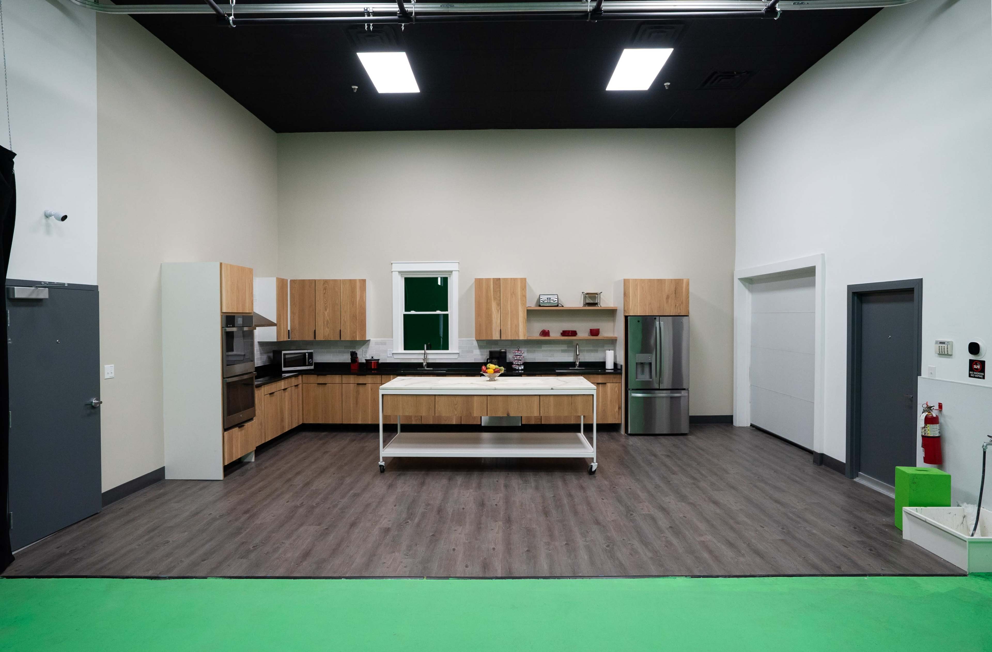 The image shows a spacious kitchen with wooden cabinetry, a central island, stainless steel appliances, and green flooring.