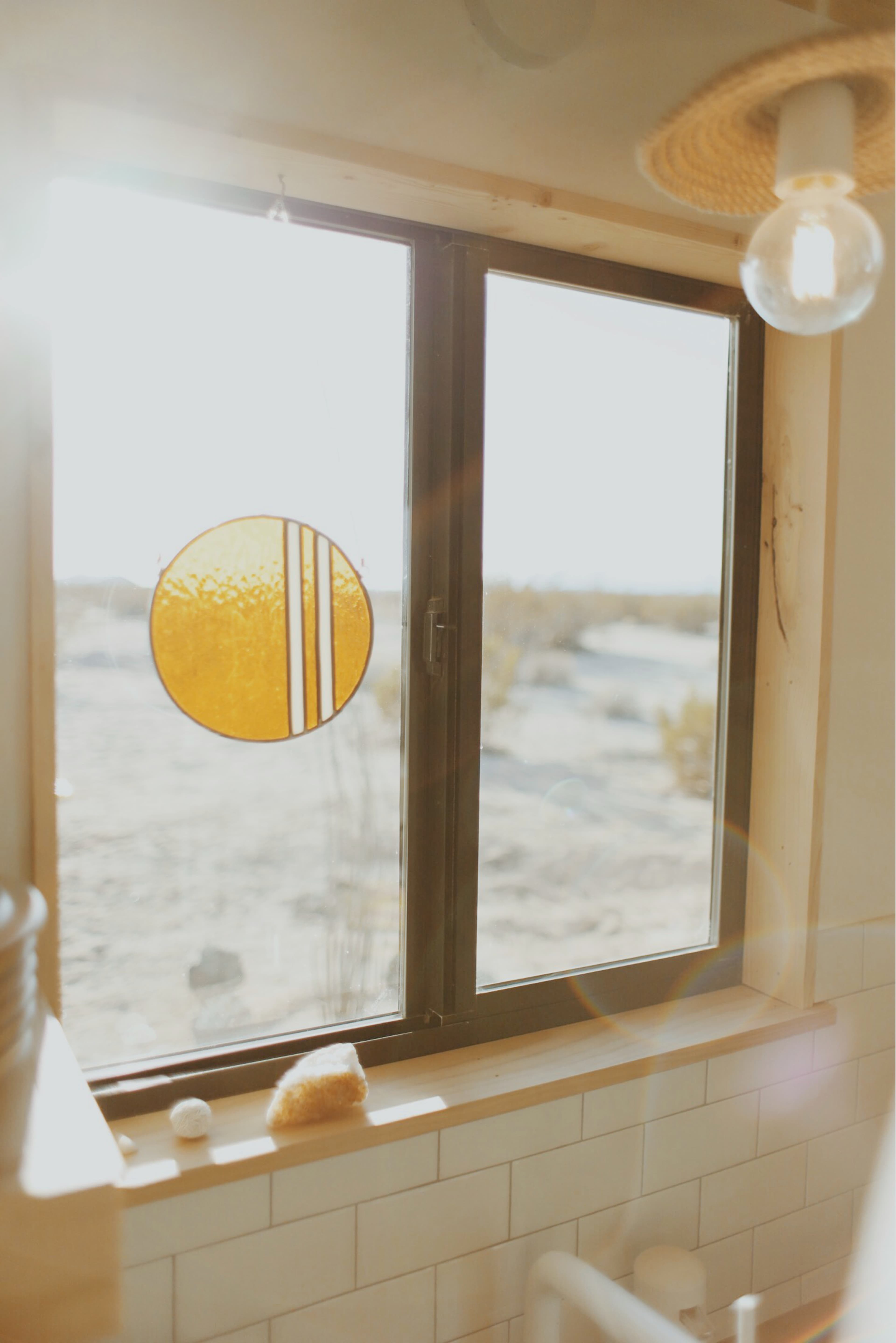 A bright sun shines through a window adorned with a round, orange stained glass hanging, illuminating a bathroom scene with desert views outside.