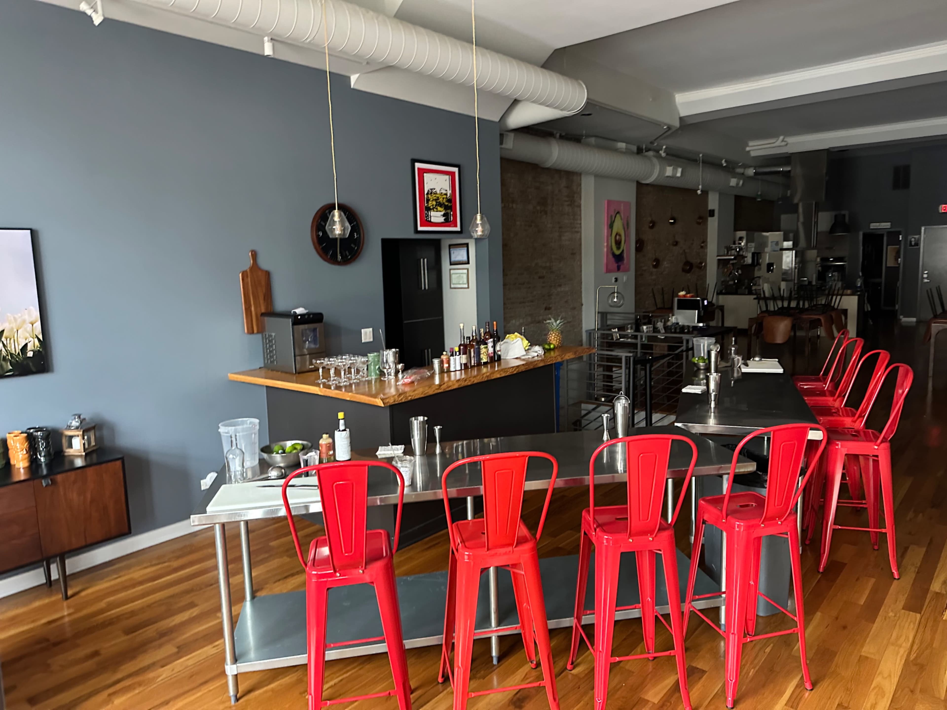 The image shows a modern bar area with red stools, a wooden countertop, and various bottles and kitchen equipment in a spacious, industrial-style setting.