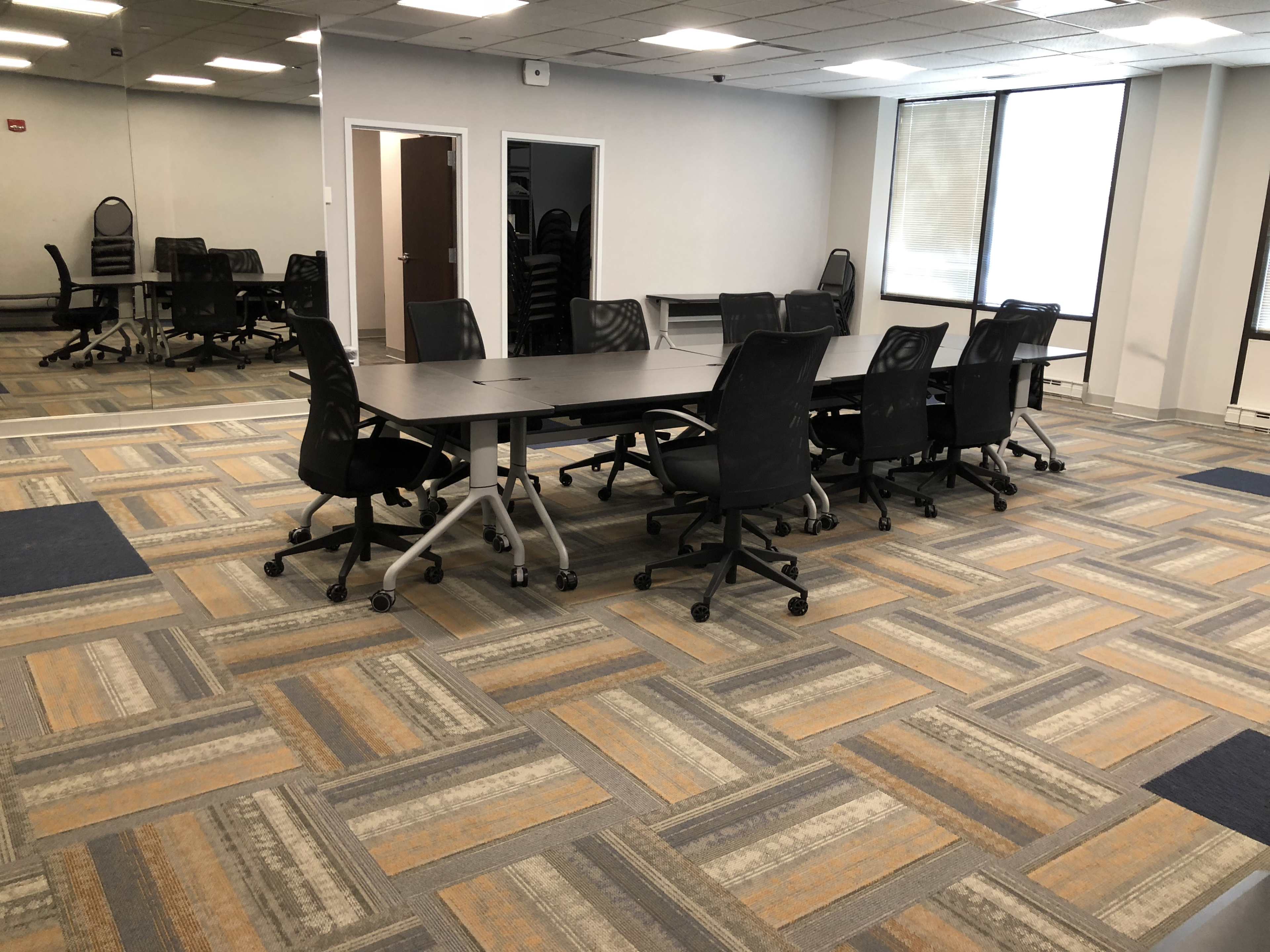 The image shows a conference room featuring a large rectangular table surrounded by black ergonomic chairs on a patterned carpet with gray and orange tones.