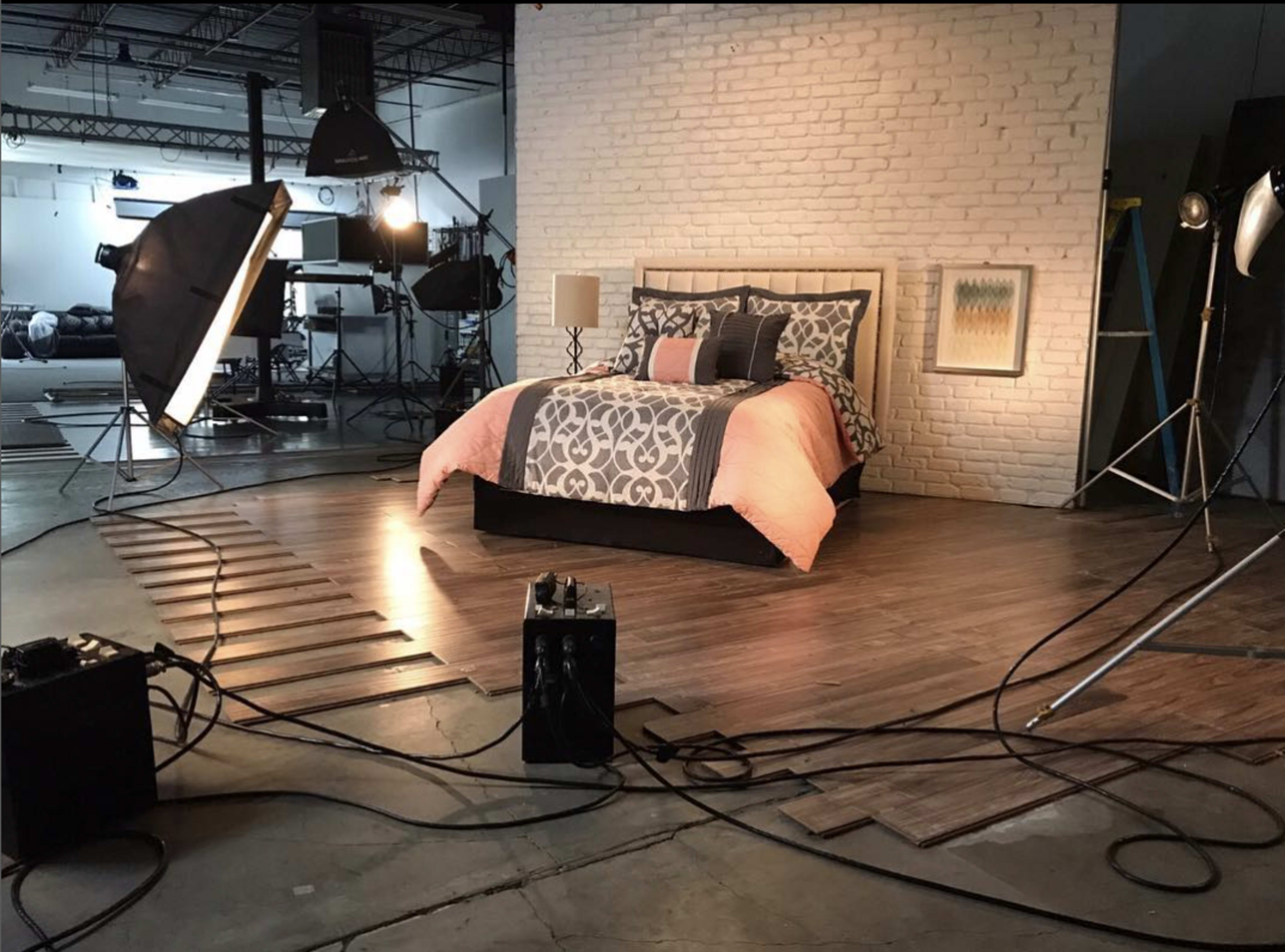 A studio setup features a bed with a patterned comforter against a brick wall, surrounded by various lights and cables on a wooden floor.