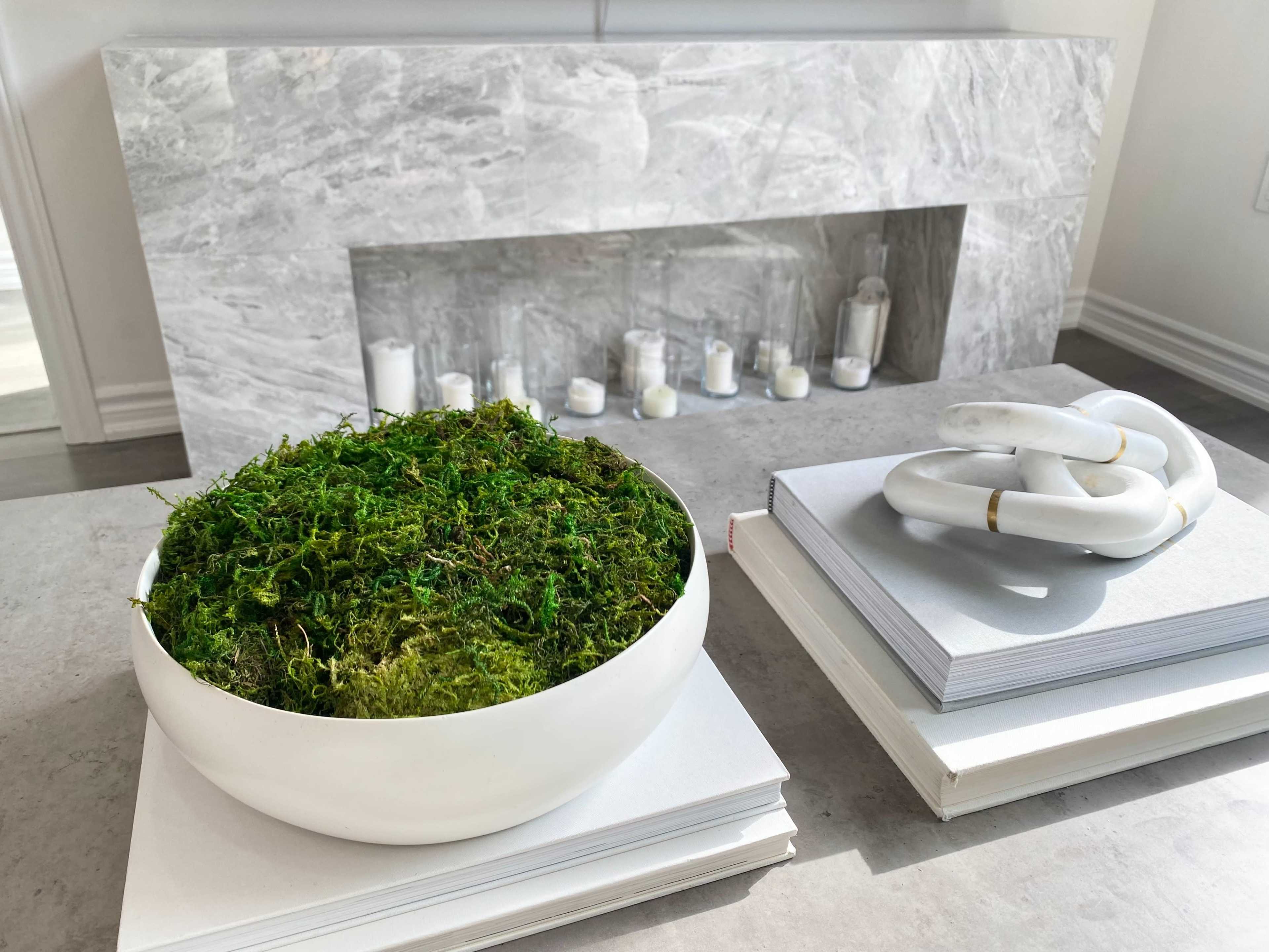 A white bowl filled with moss sits on a stack of gray books next to a marble fireplace with white candles arranged on the ledge.
