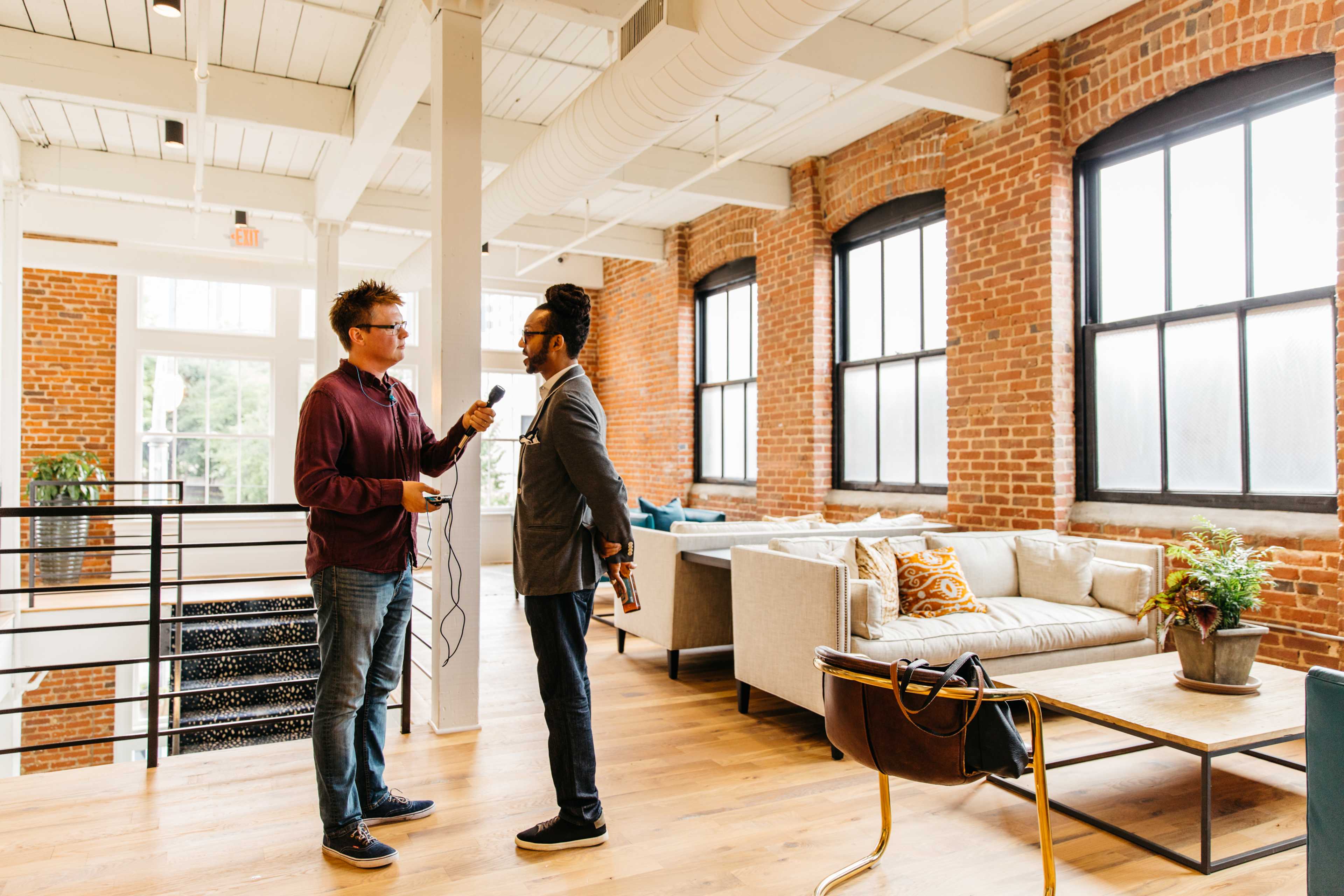 A man interviews another man in a bright, stylish loft with large windows and brick walls.