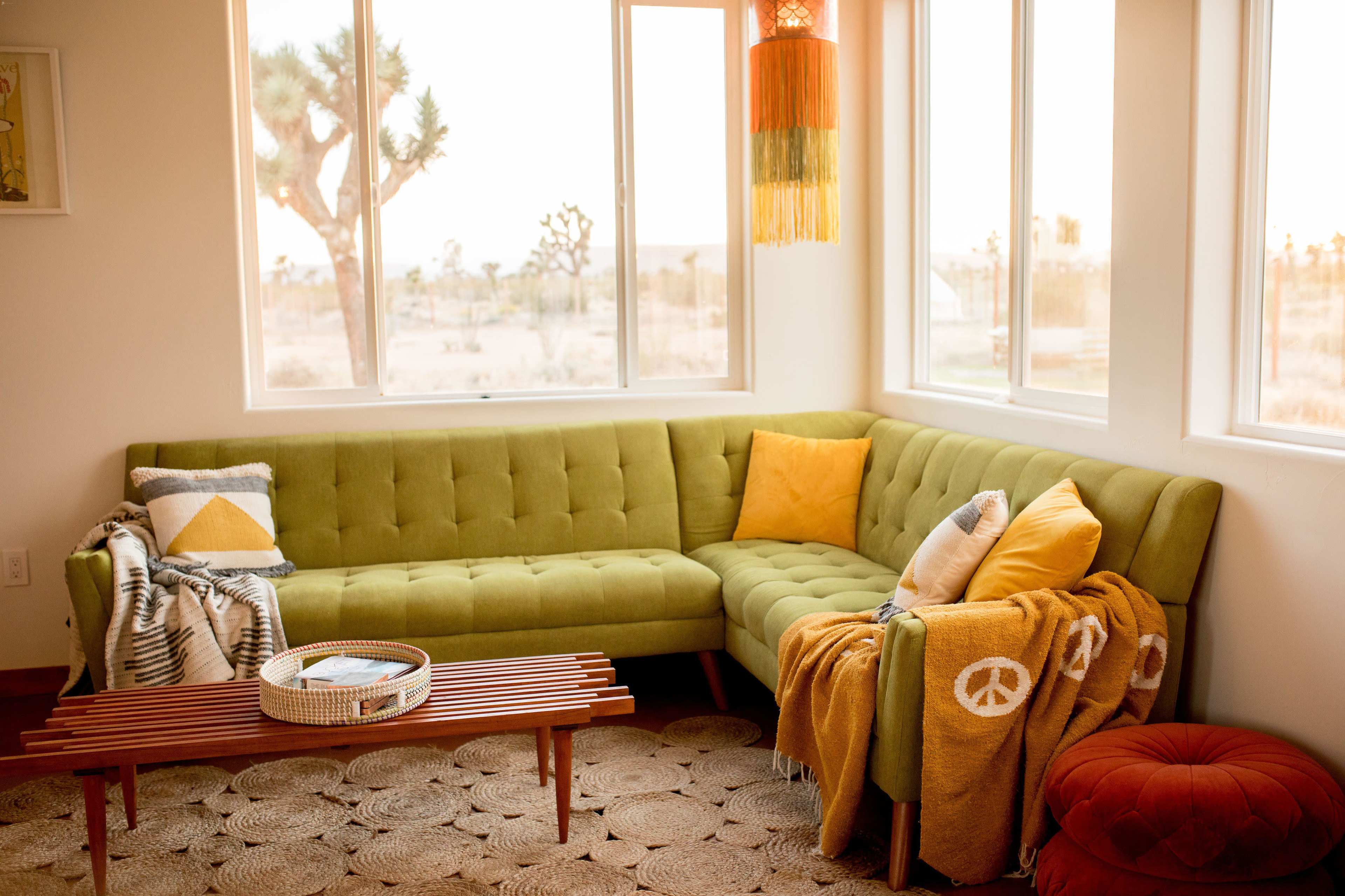 A cozy living room with a green sectional sofa, yellow pillows, a wooden coffee table, and a bright window overlooking a desert landscape.