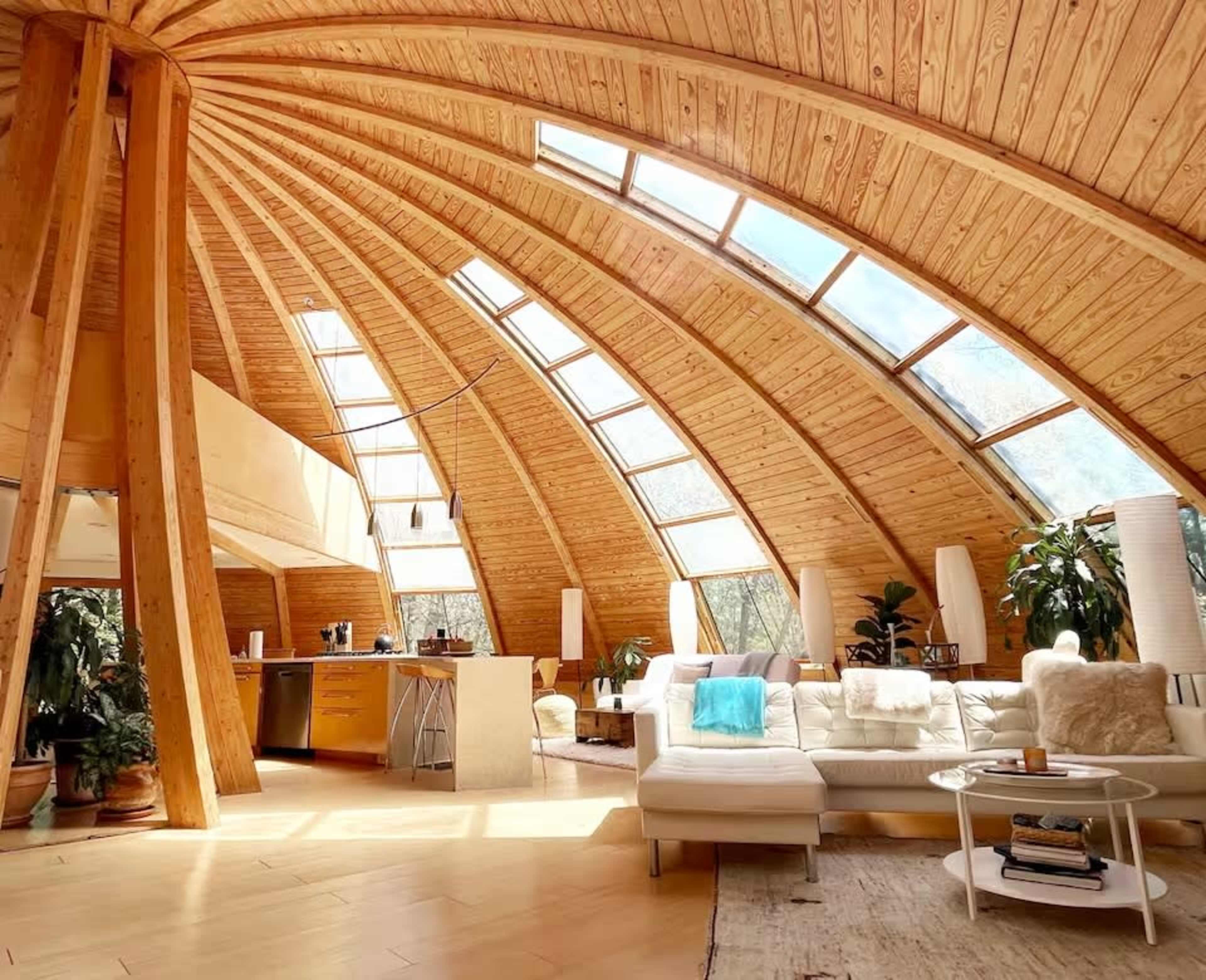 The image shows a modern, wooden interior of a dome-shaped living space with large windows and a cozy seating area.