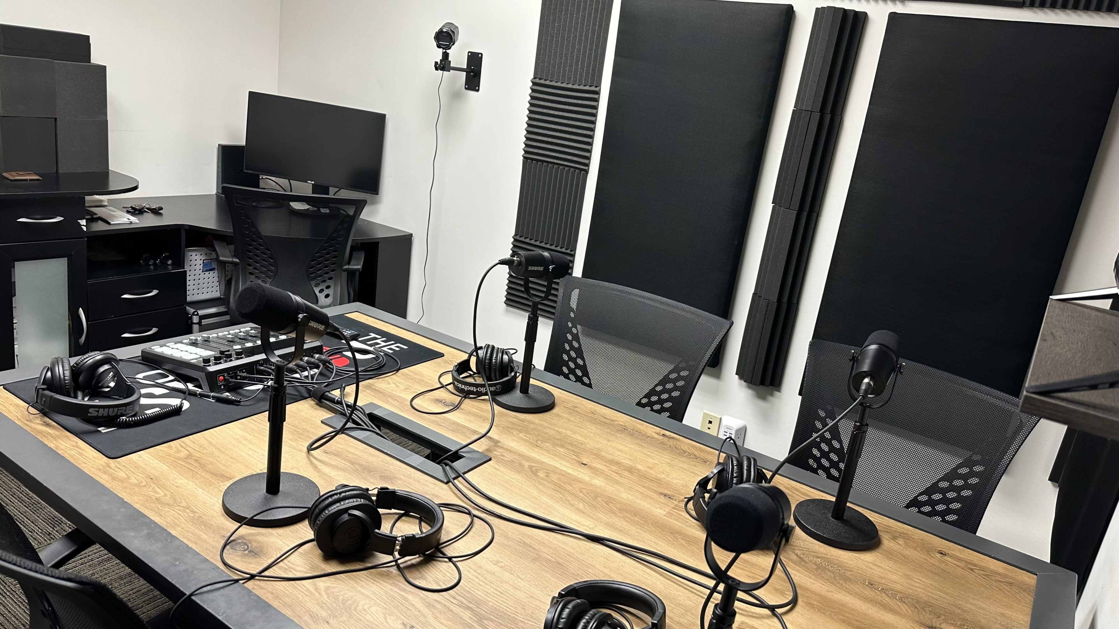 The image shows a recording studio equipped with a wooden table, multiple microphones, headphones, and soundproofing panels on the walls.
