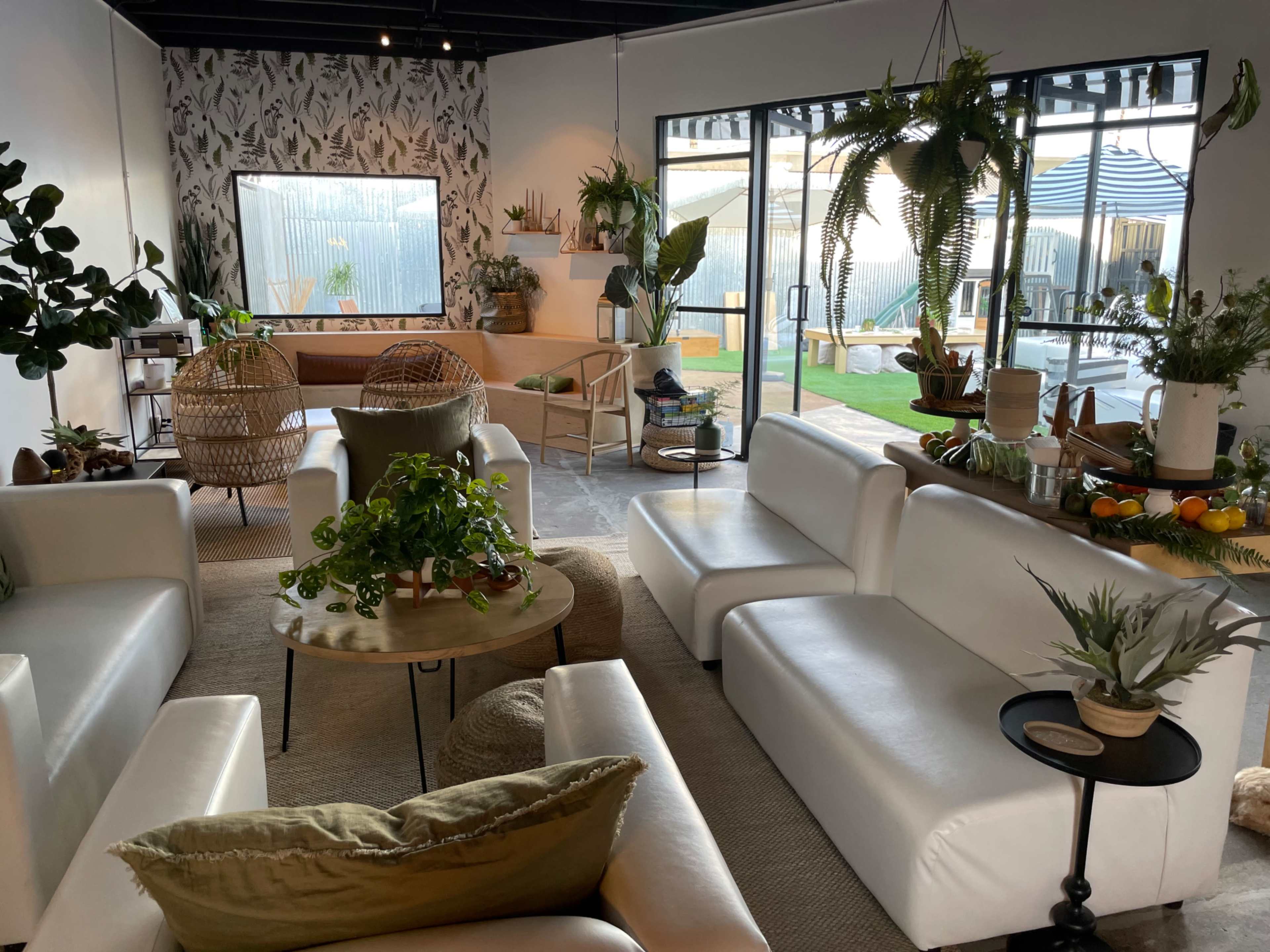 The image shows a modern, well-lit indoor space with white sofas, a round wooden table, various plants, and a view of an outdoor area through large glass doors.