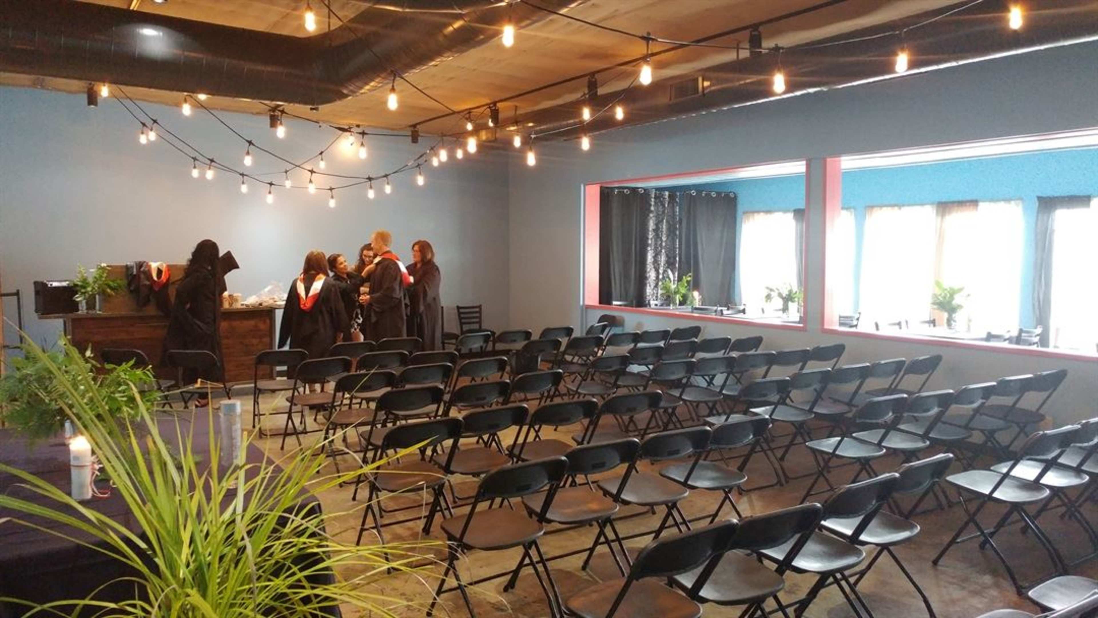 A spacious room is arranged with rows of black chairs facing a snacks table, while several individuals in graduation robes converse near the back.