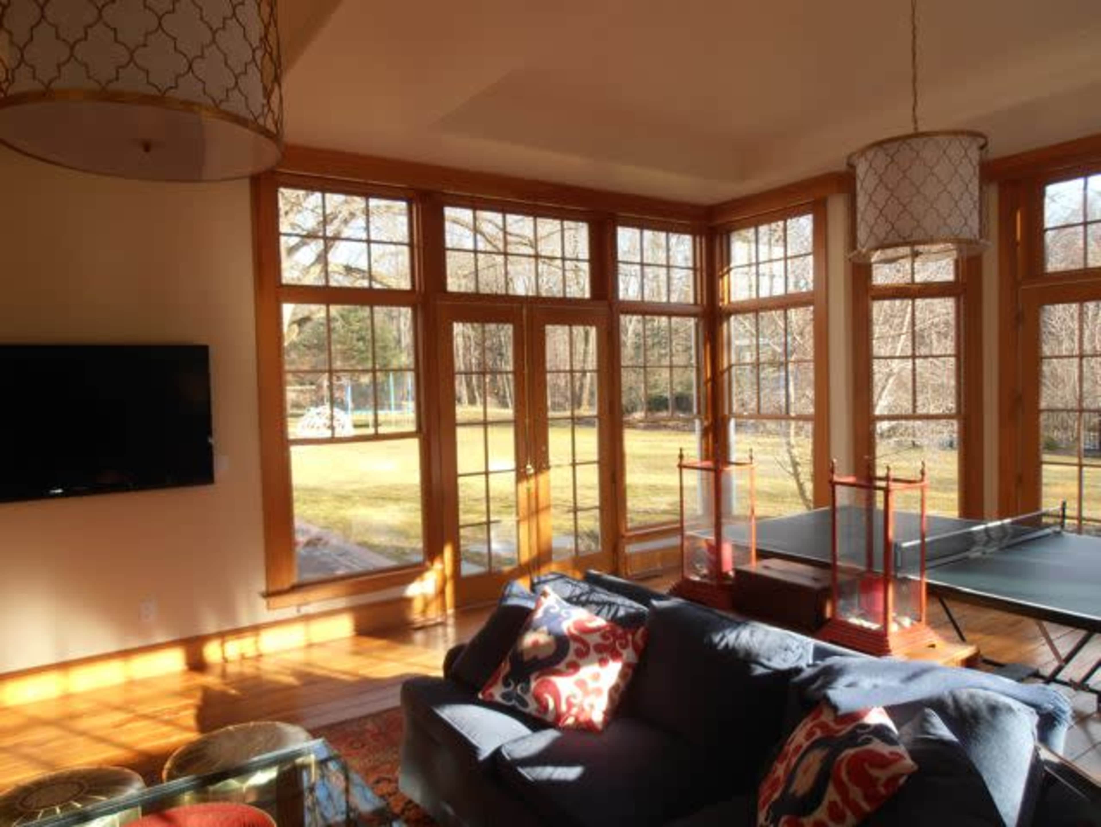 A bright living room features large windows with a view of a grassy yard, a sofa with patterned cushions, and a table tennis setup.