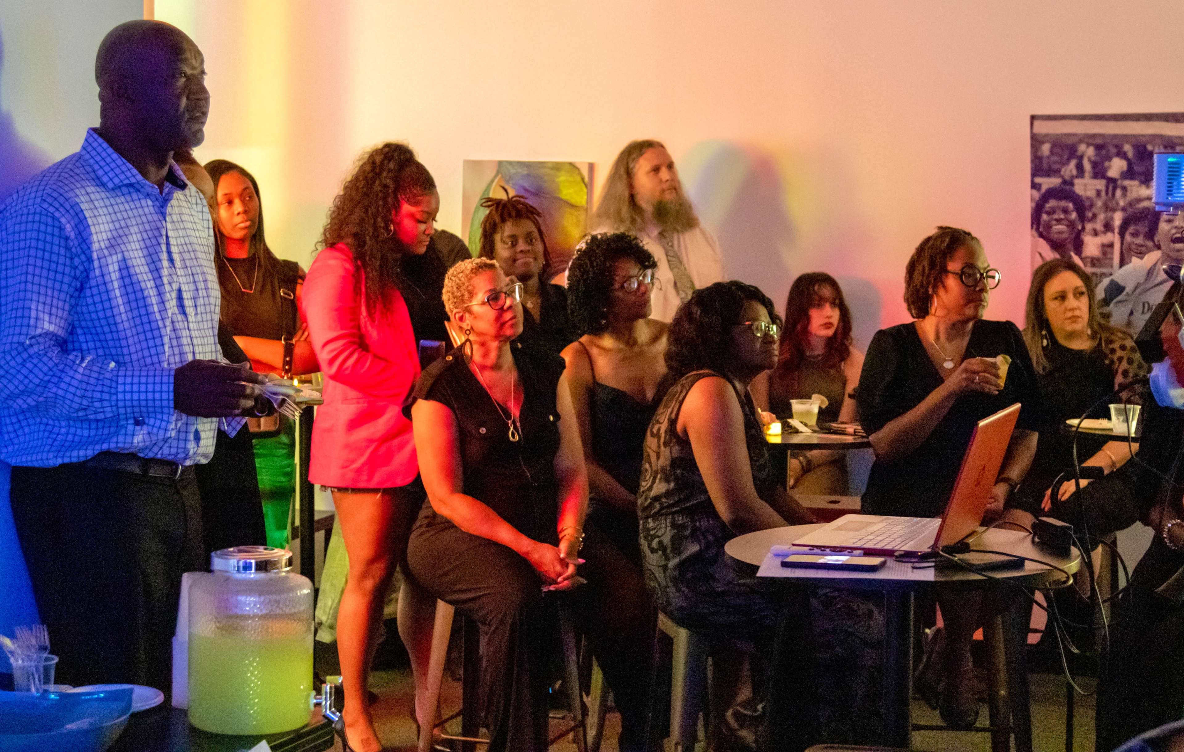 A diverse group of people attentively watches a presentation in a brightly lit room with colorful lighting.