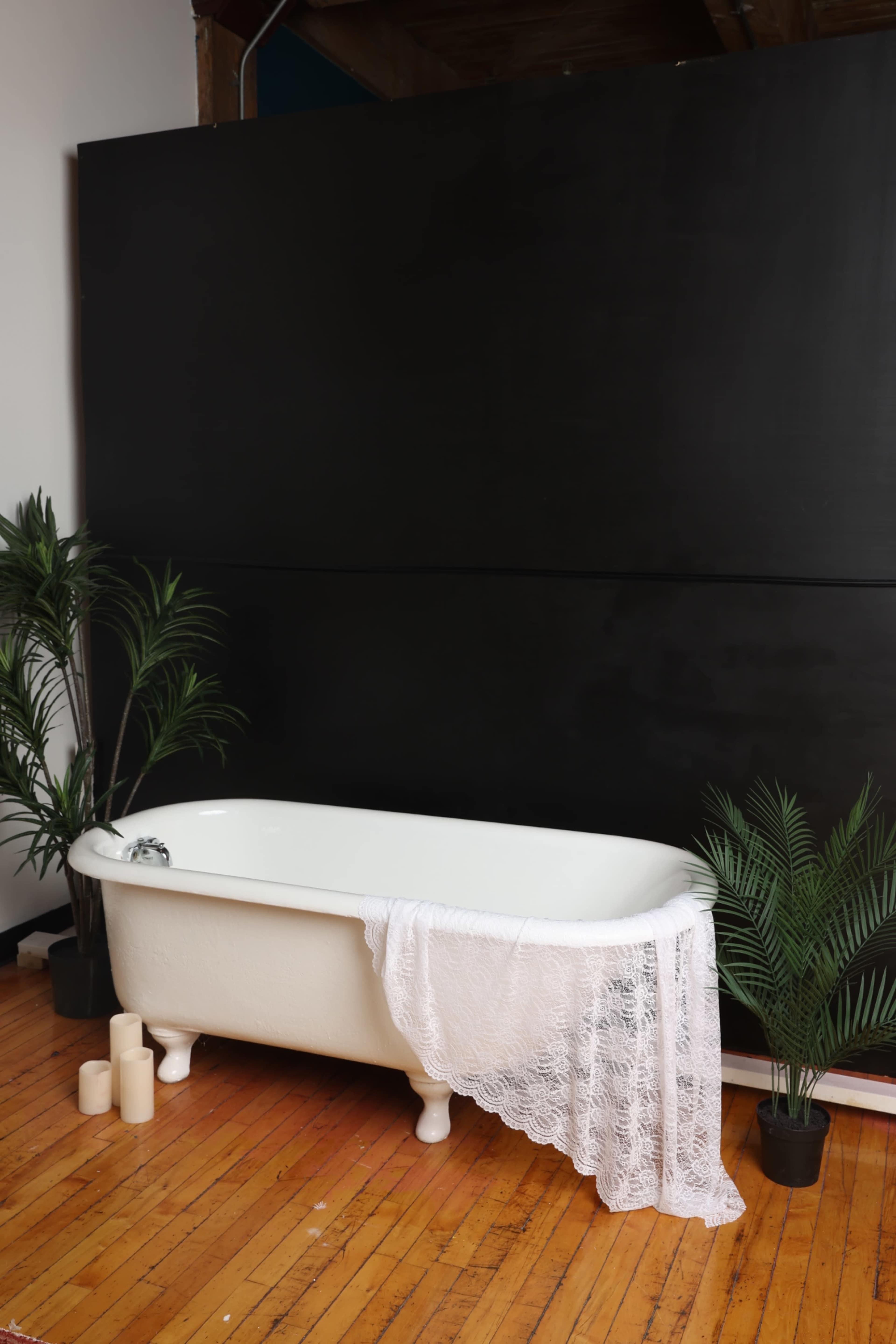 A white freestanding bathtub with a lace cloth draped over it is surrounded by potted plants on a wooden floor against a black wall.