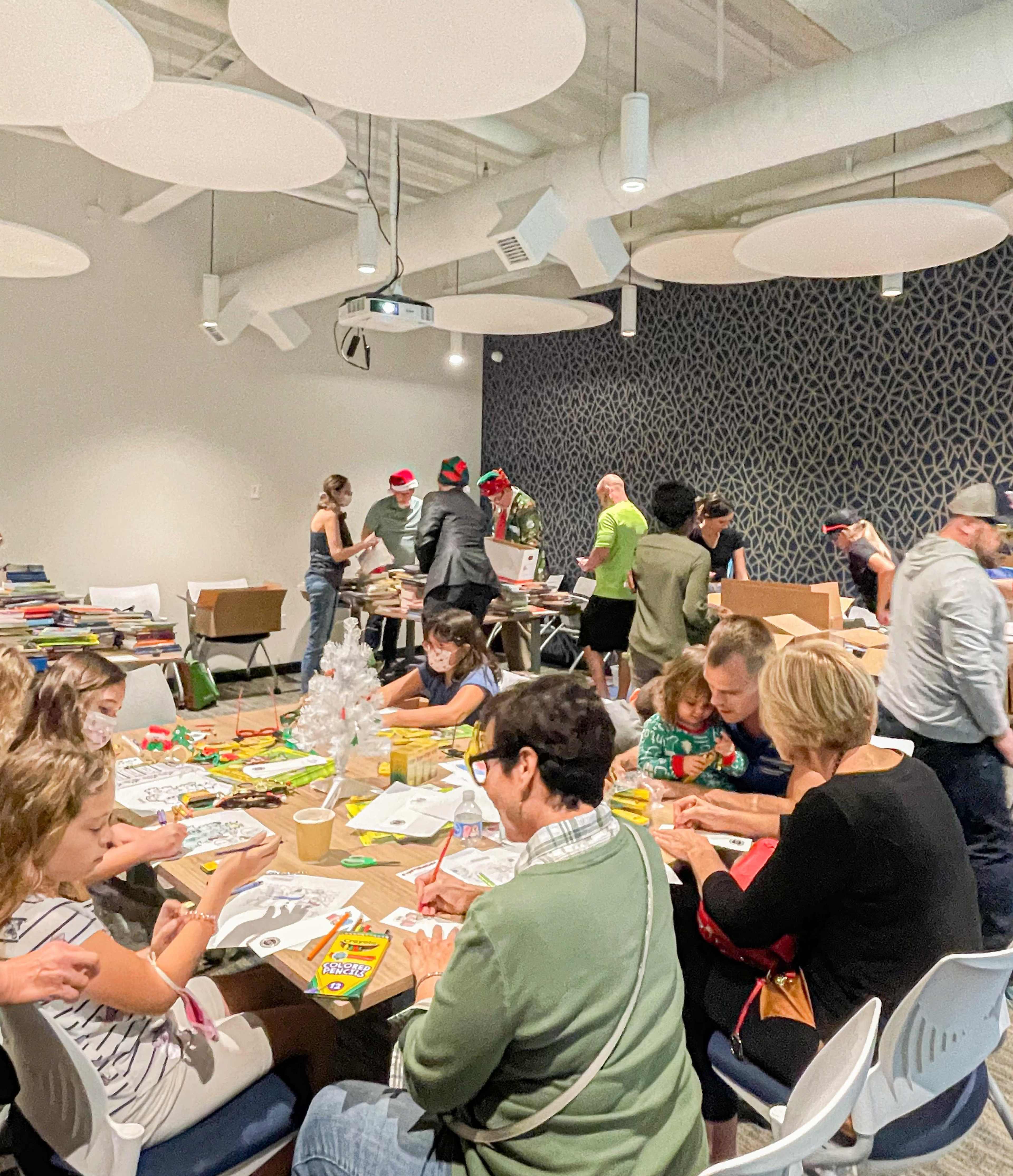 A group of people, including children and adults, are engaged in arts and crafts activities around a table in a well-lit room decorated for a holiday gathering.
