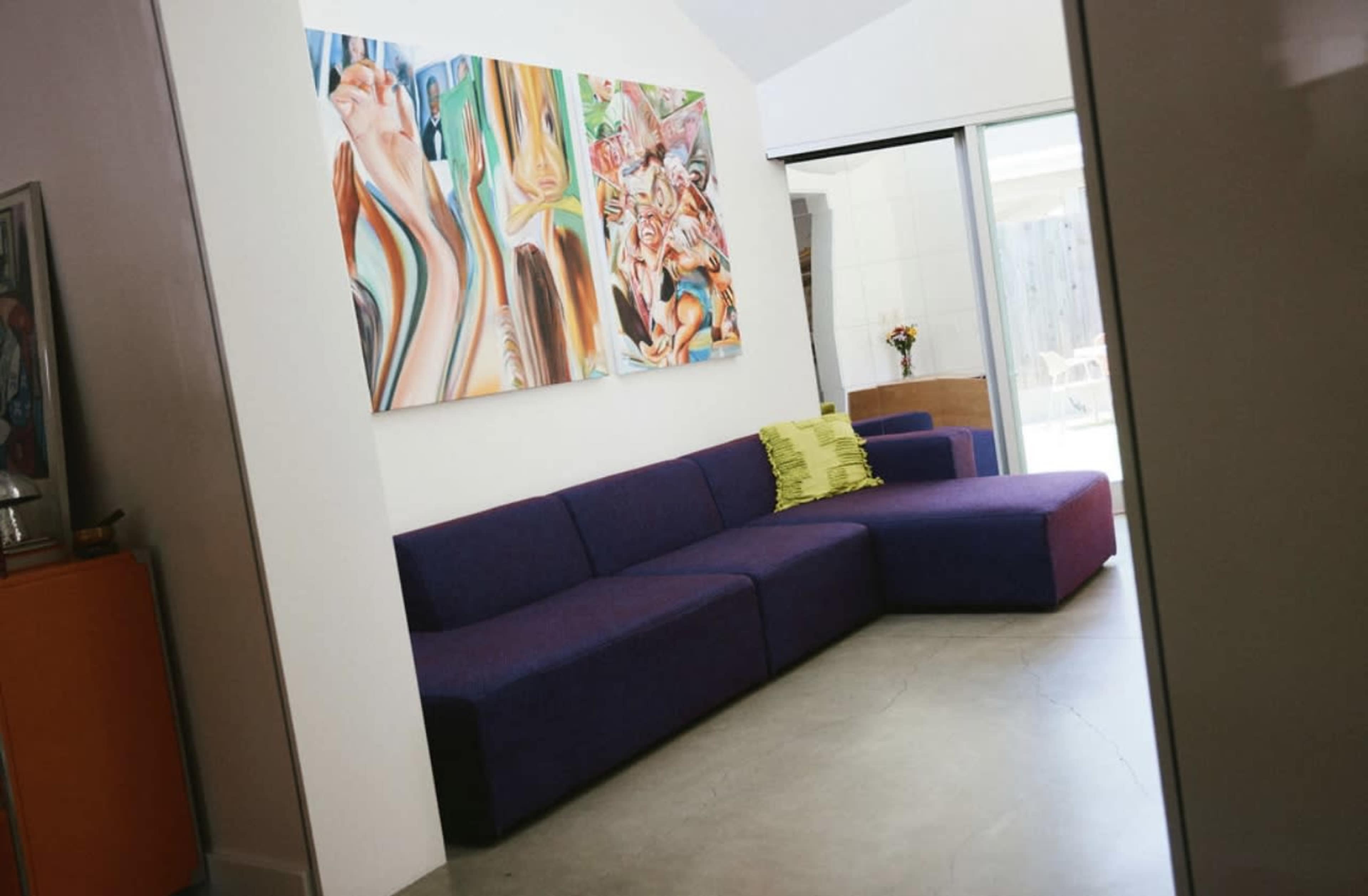 A modern living room with a purple sectional sofa against a white wall adorned with colorful abstract paintings.