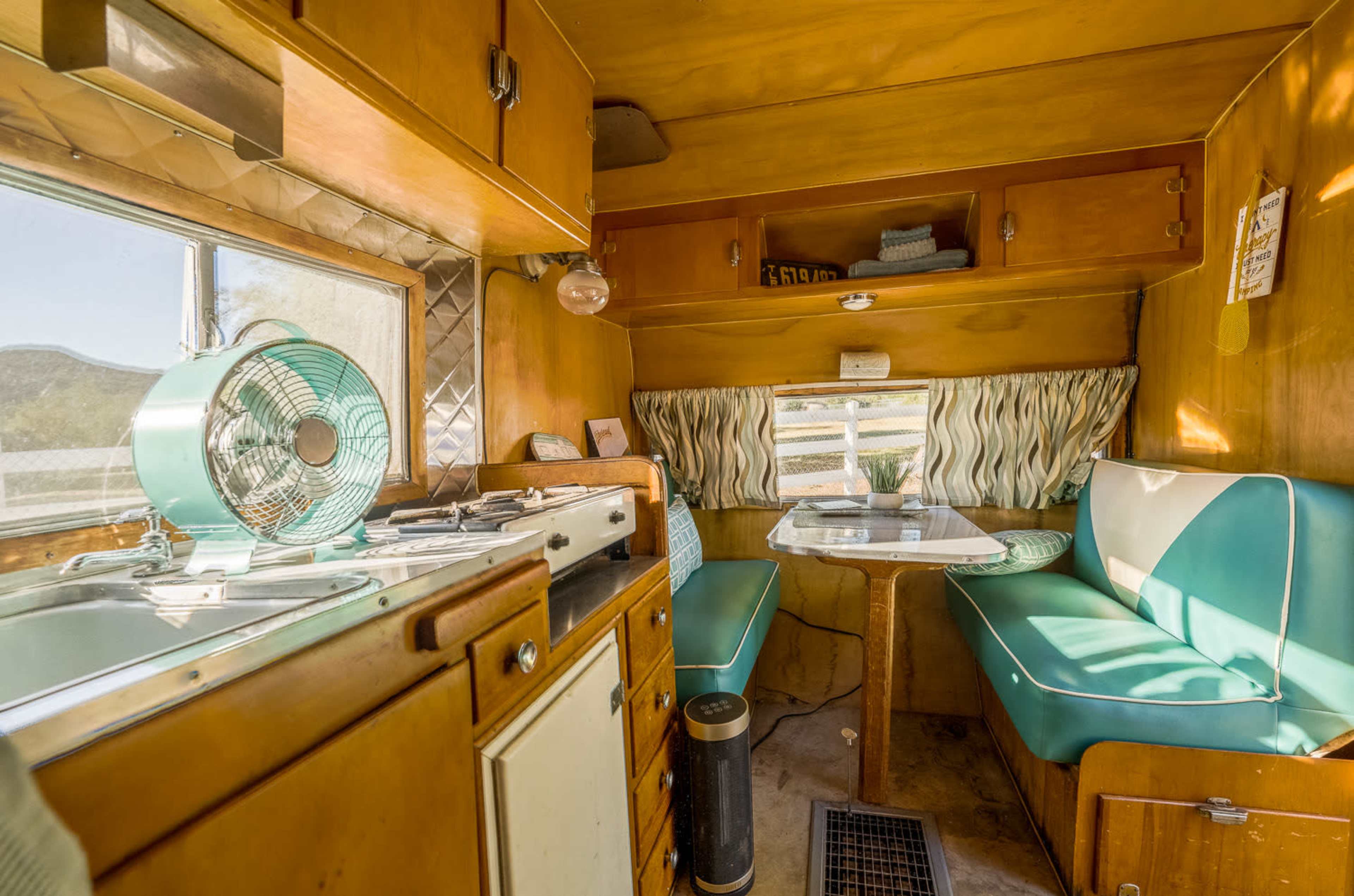 The interior of a vintage camper features a kitchenette with a stove, a table for dining, and turquoise and white seating.