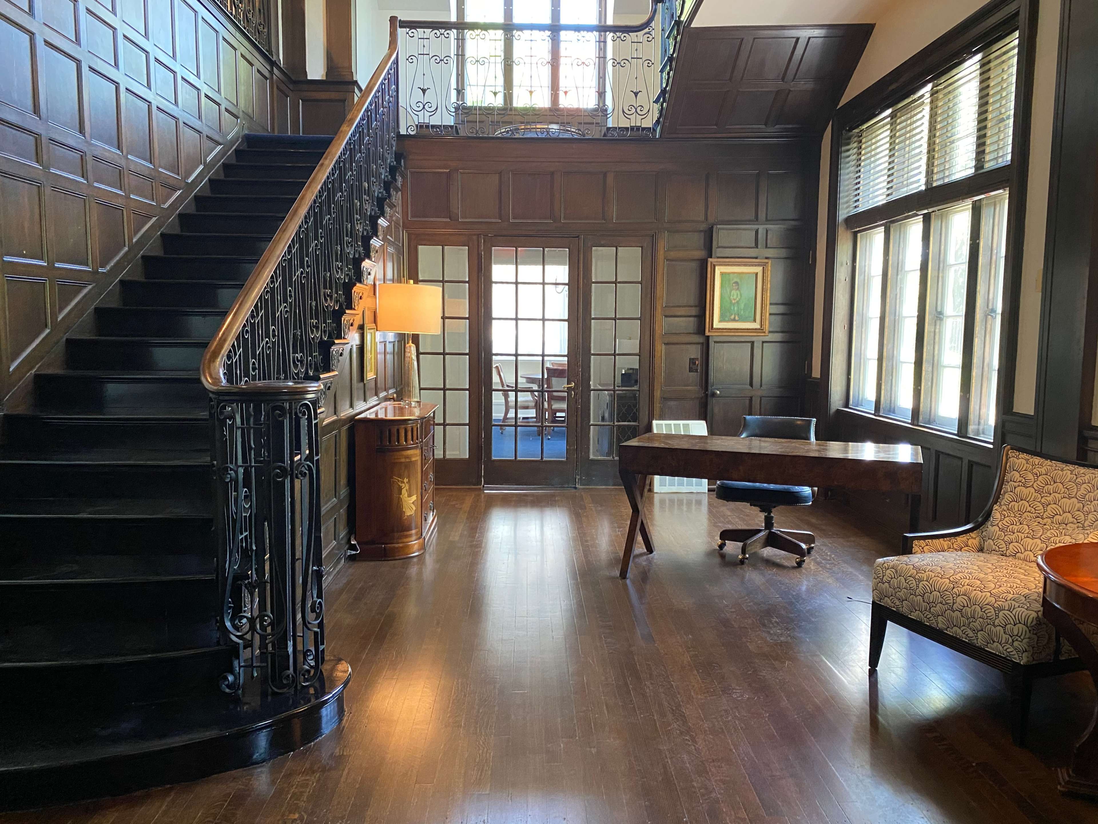 The image shows a spacious interior featuring a wooden staircase, a desk, a chair, and large windows that brighten the room.