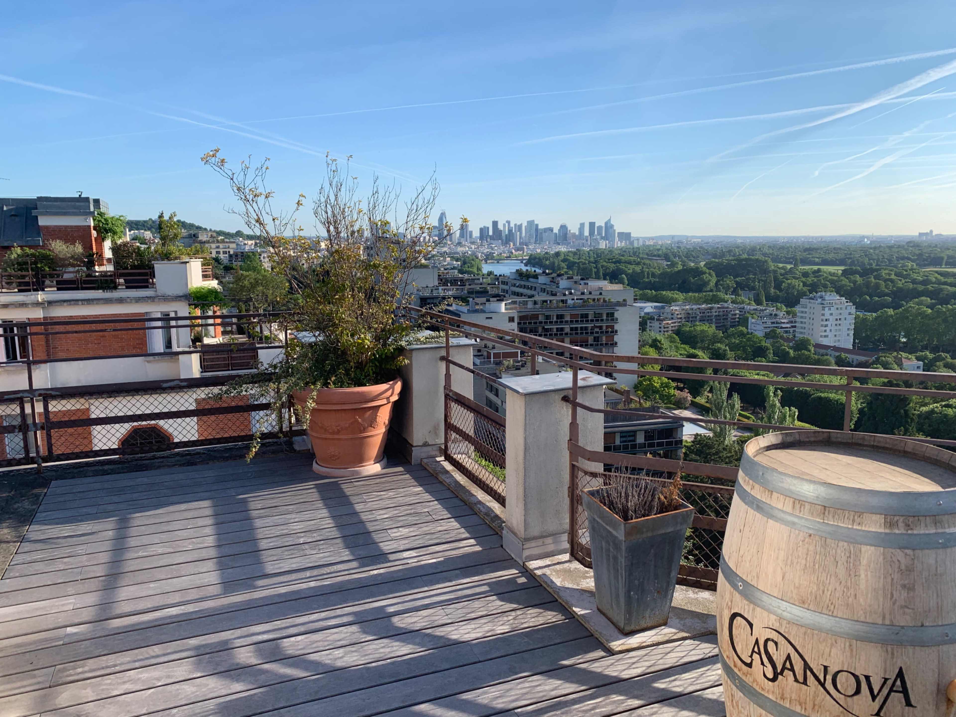 A rooftop terrace with wooden flooring overlooks a city skyline amid greenery and a large planter.