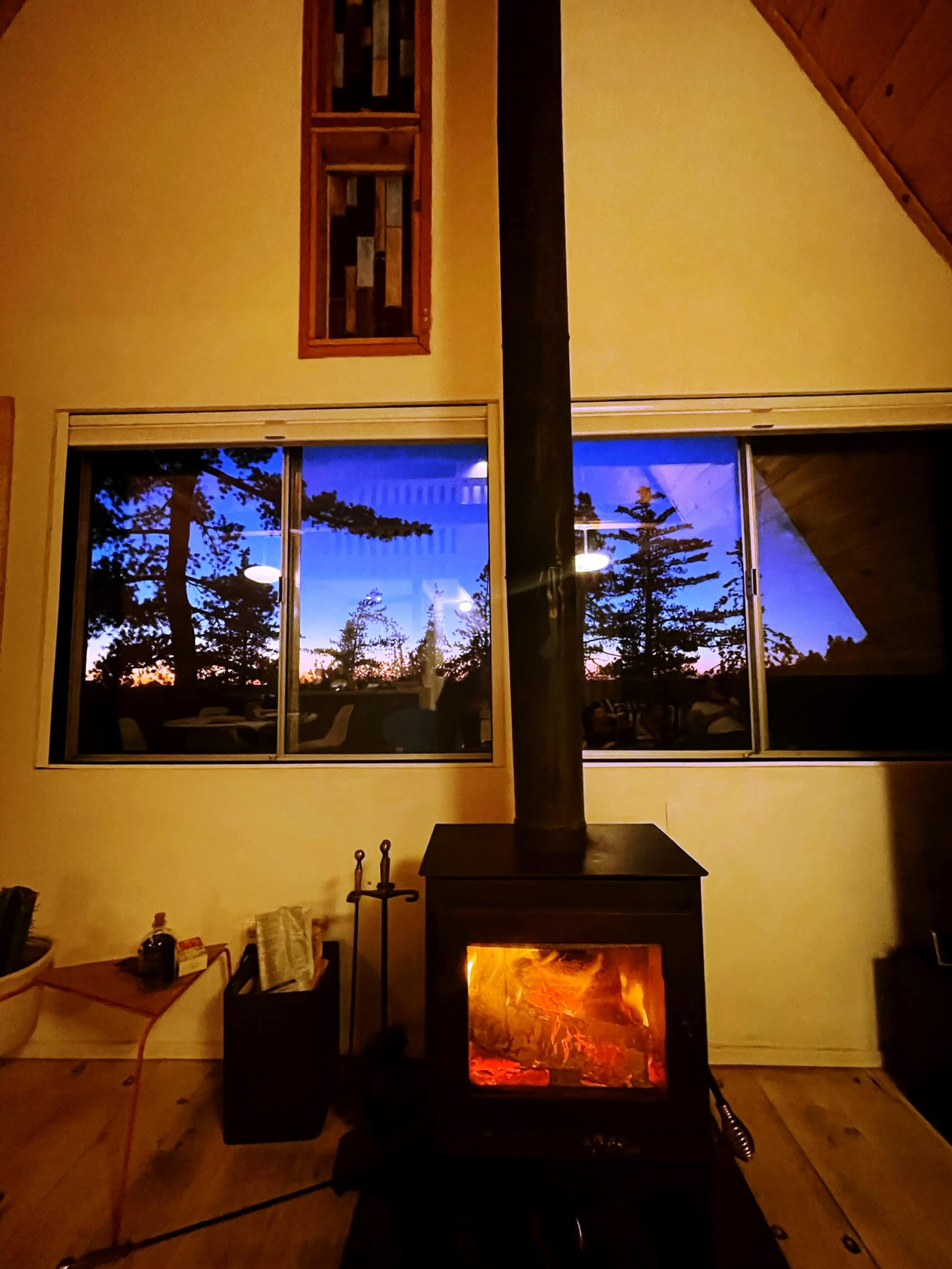 A wood-burning stove emits light in a cabin, reflecting the twilight sky and trees in the large window behind it.