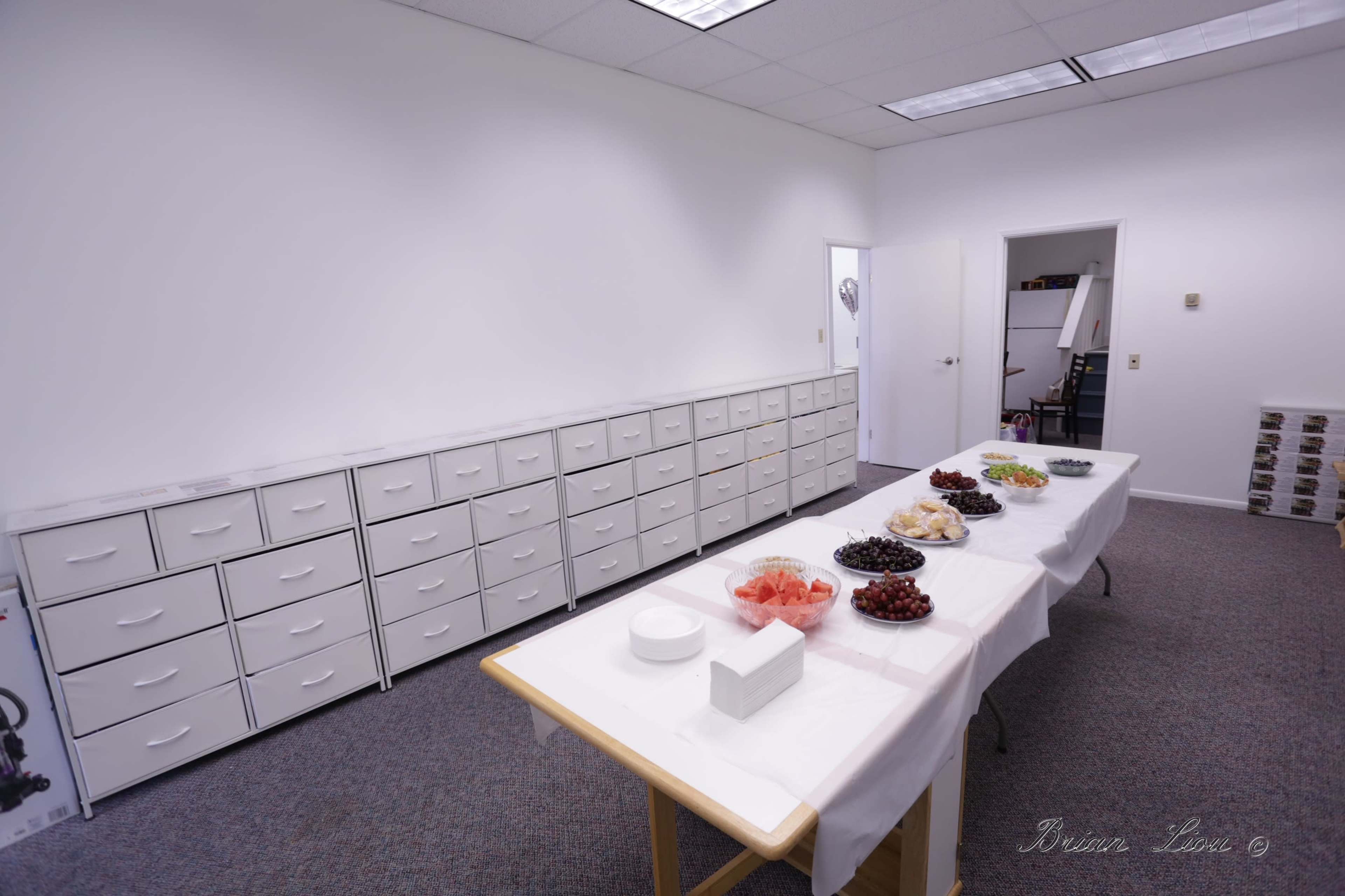 A spacious room with white walls, featuring long tables set with various bowls of fruit and a row of cabinets lining one side.