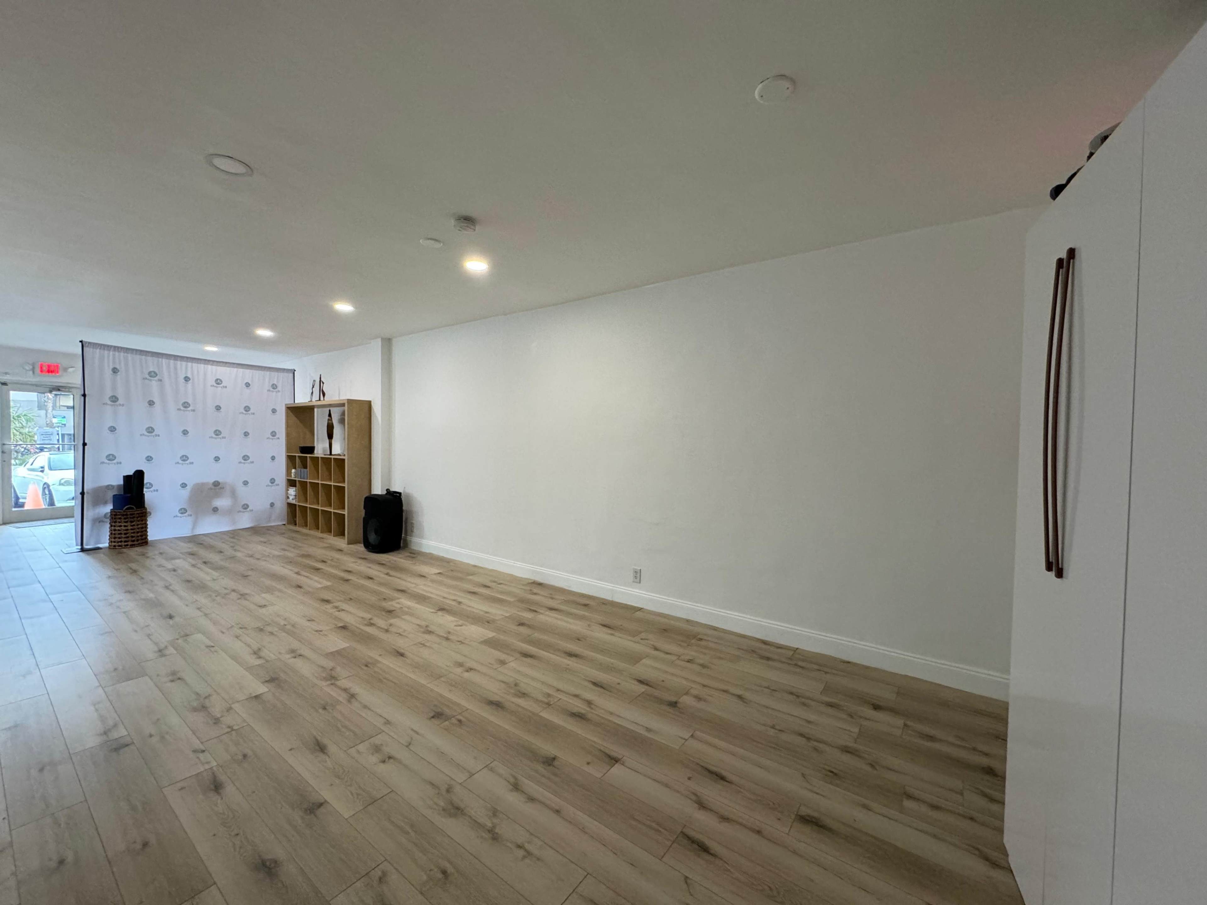 A spacious, empty room with light-colored wooden flooring and a white wall featuring a refrigerator and a shelf against one side.