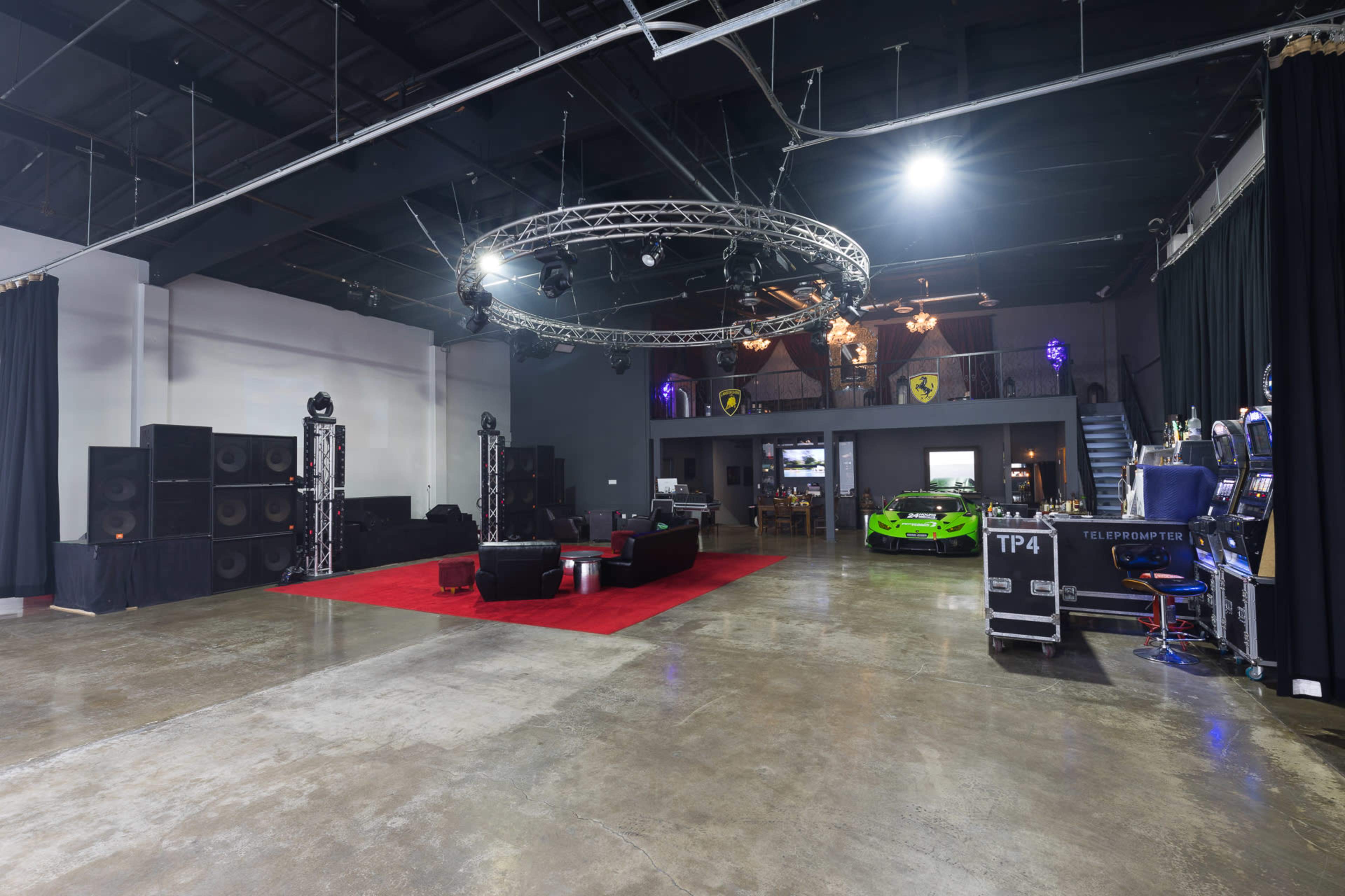 The spacious interior features a polished concrete floor, stage lighting, seating area with black couches, a green sports car, and entertainment equipment.