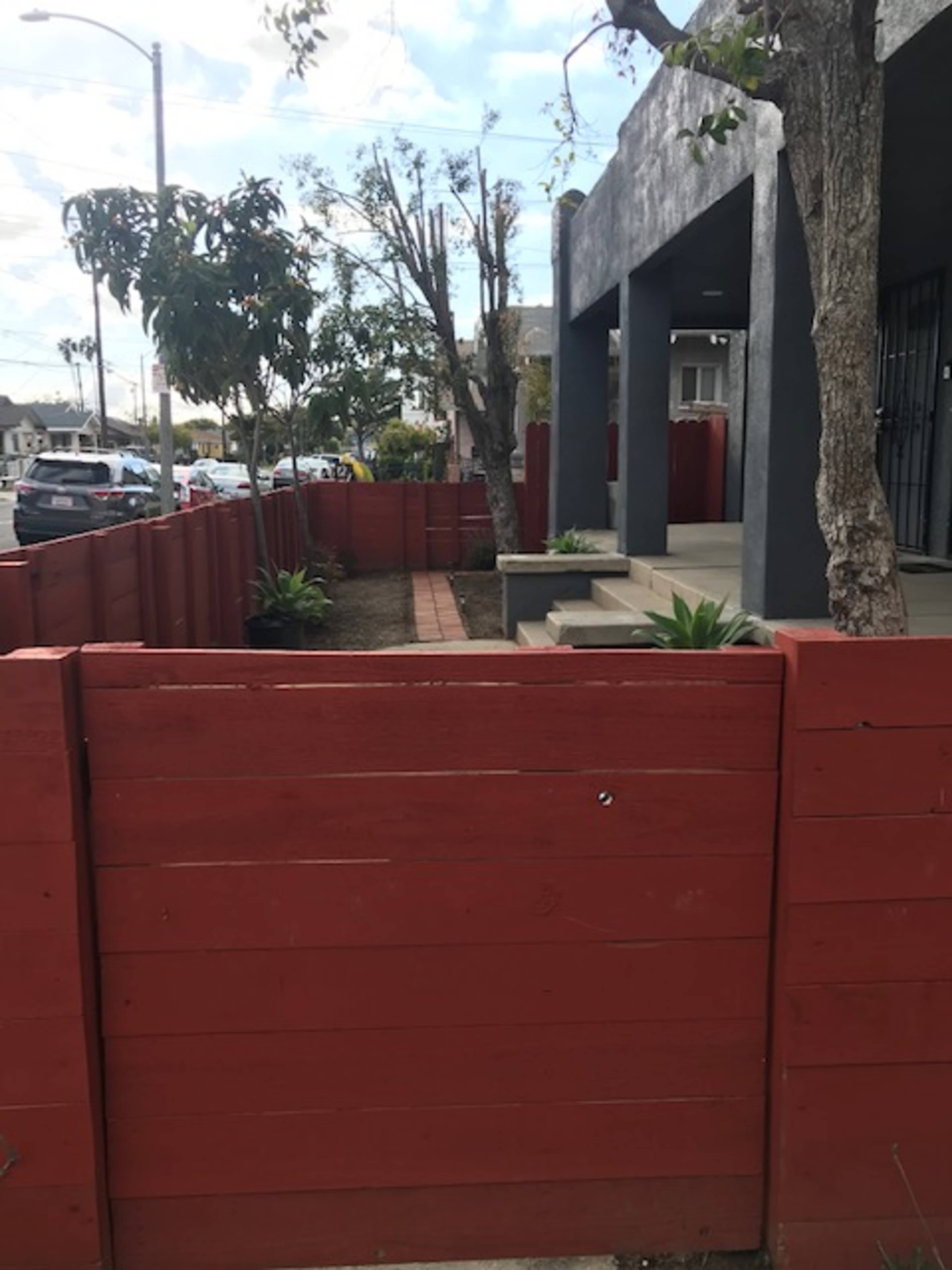 A red fenced area with potted plants and a walkway next to a building, set against a partly cloudy sky.