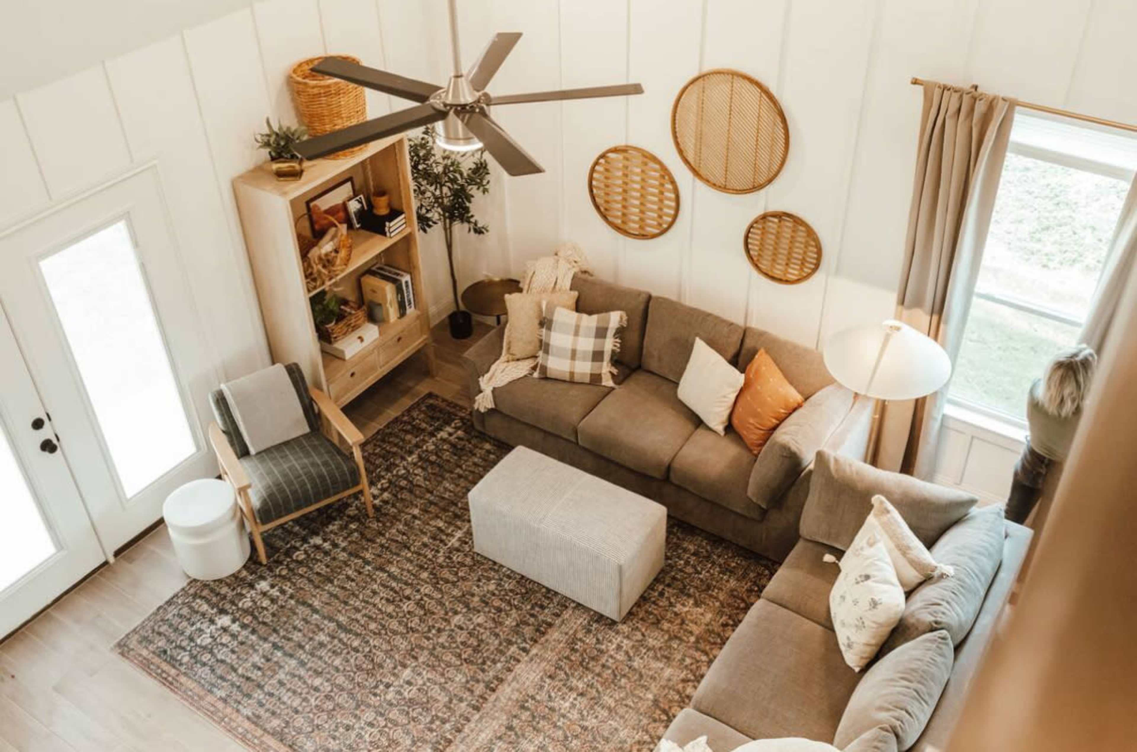 A spacious living room features a sectional sofa, a coffee table, bookshelves, and decorative wall hangings, with large windows allowing natural light.