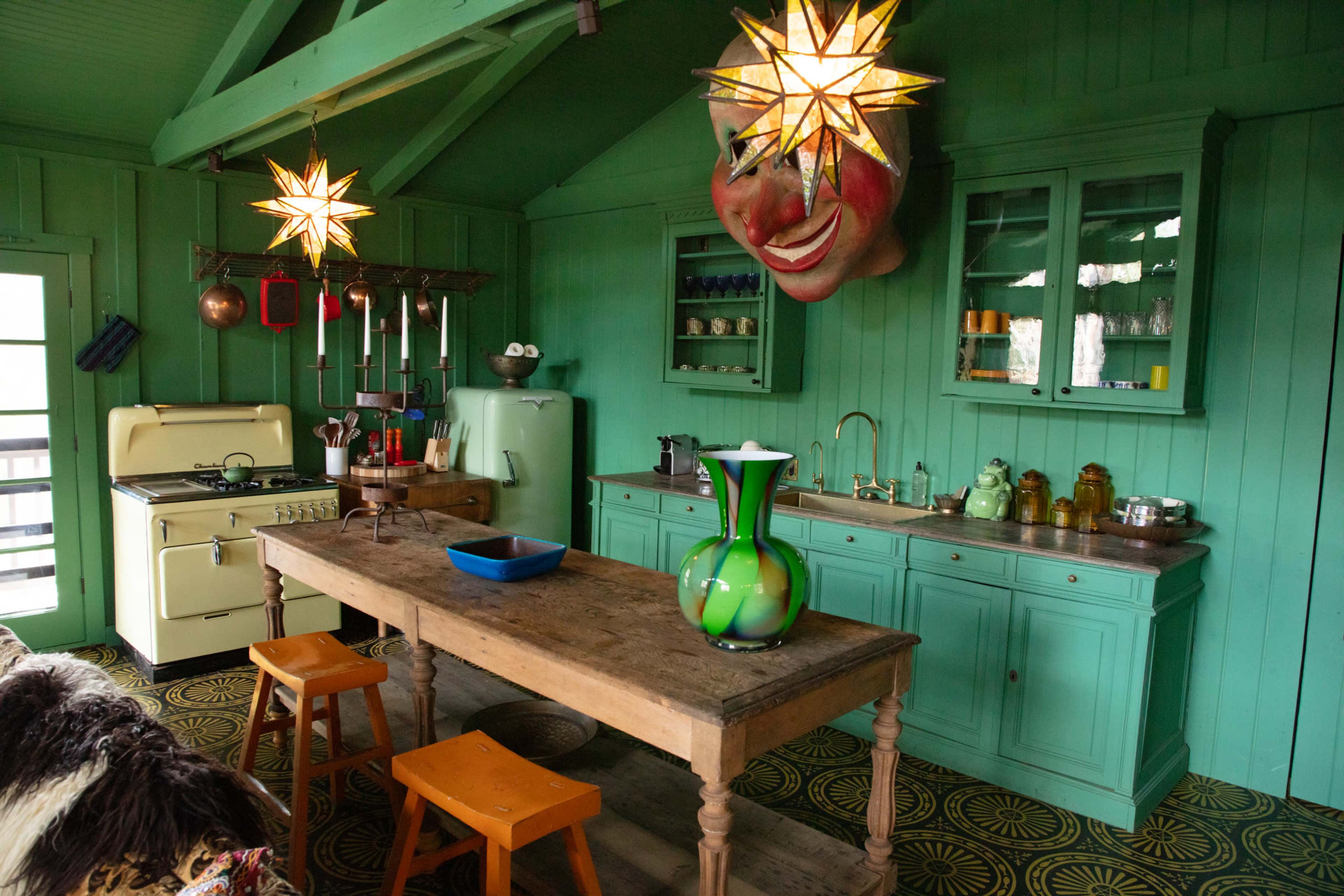 The image shows a brightly colored kitchen with turquoise walls, wooden furniture, and unique decorative elements like star-shaped light fixtures and a large mask on the wall.