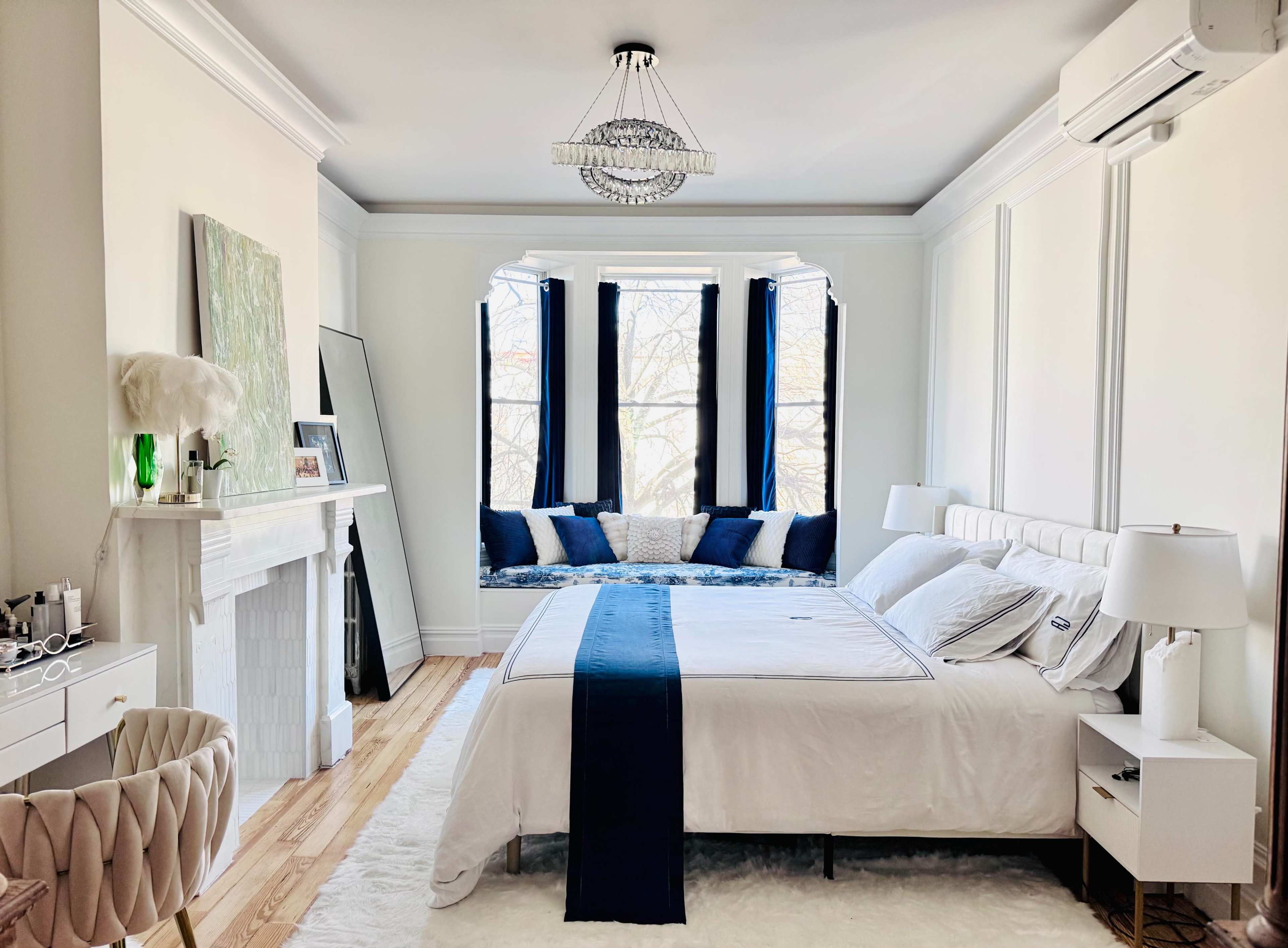 A spacious bedroom features a white bed with navy bedding, large windows with seating, and a decorative chandelier.