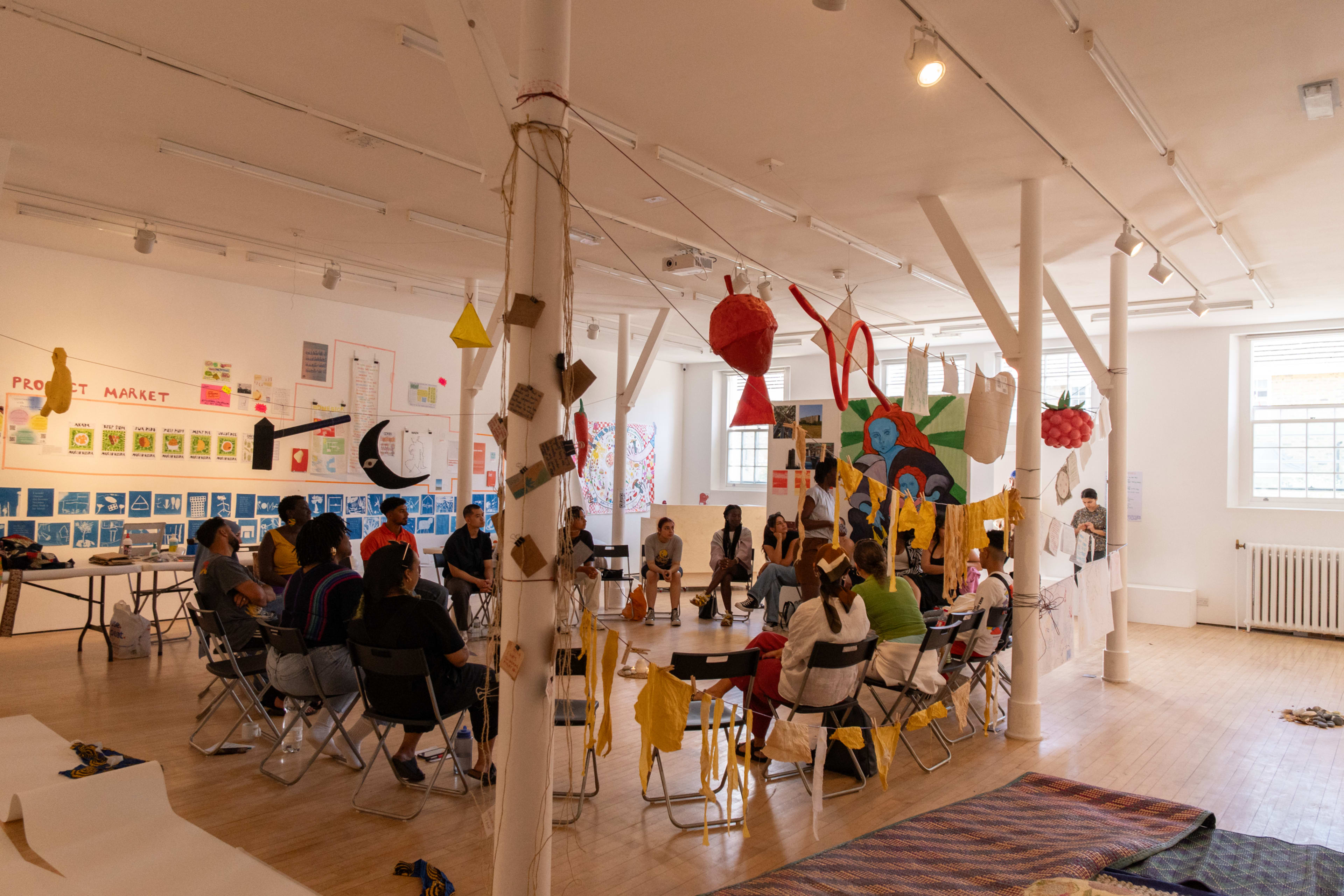 A group of people sits in a circle in a well-lit art space adorned with colorful decorations and various artworks on the walls.