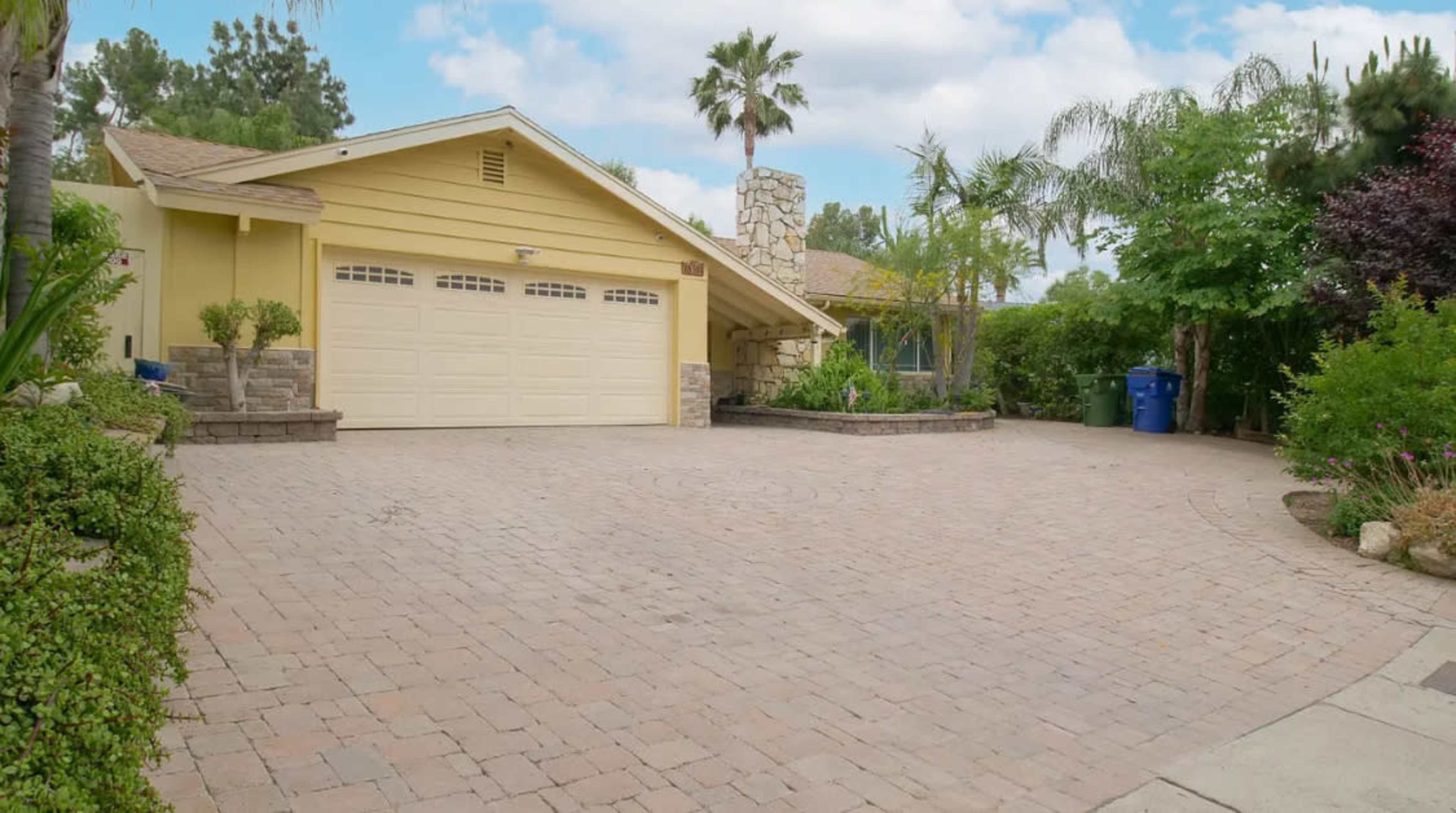 A yellow house with a stone chimney is situated next to a paved driveway and surrounded by greenery.