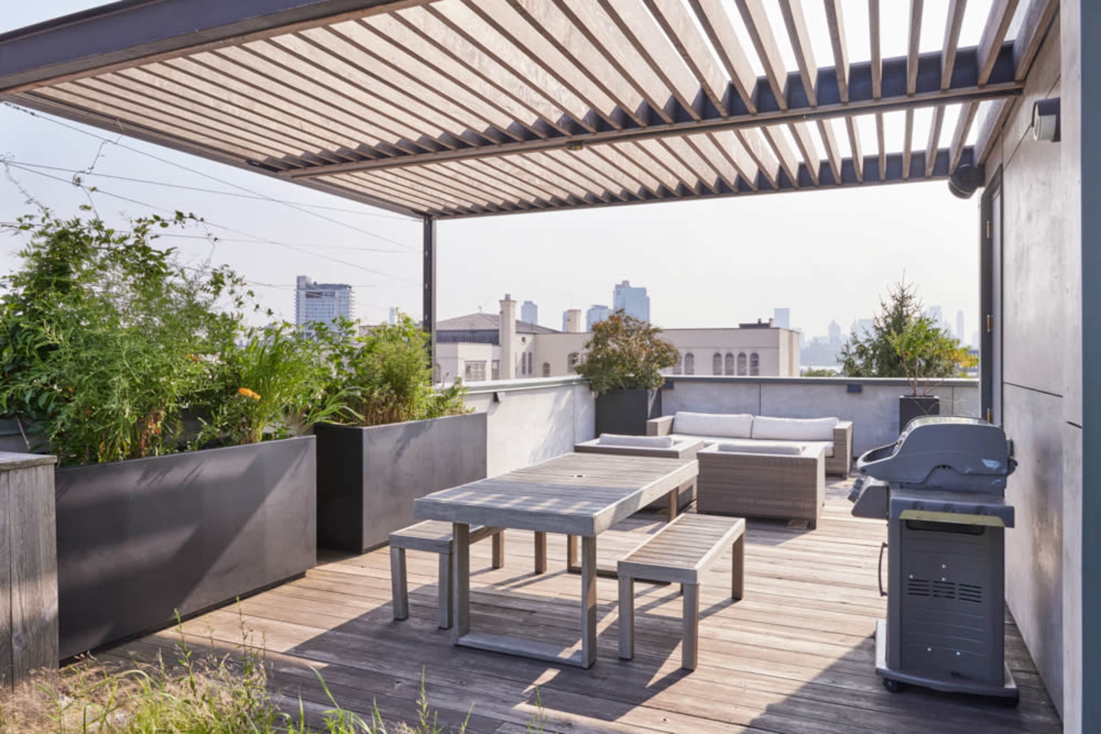 The rooftop terrace features a wooden deck, a seating area, a dining table, potted plants, and a grill under a slatted pergola.