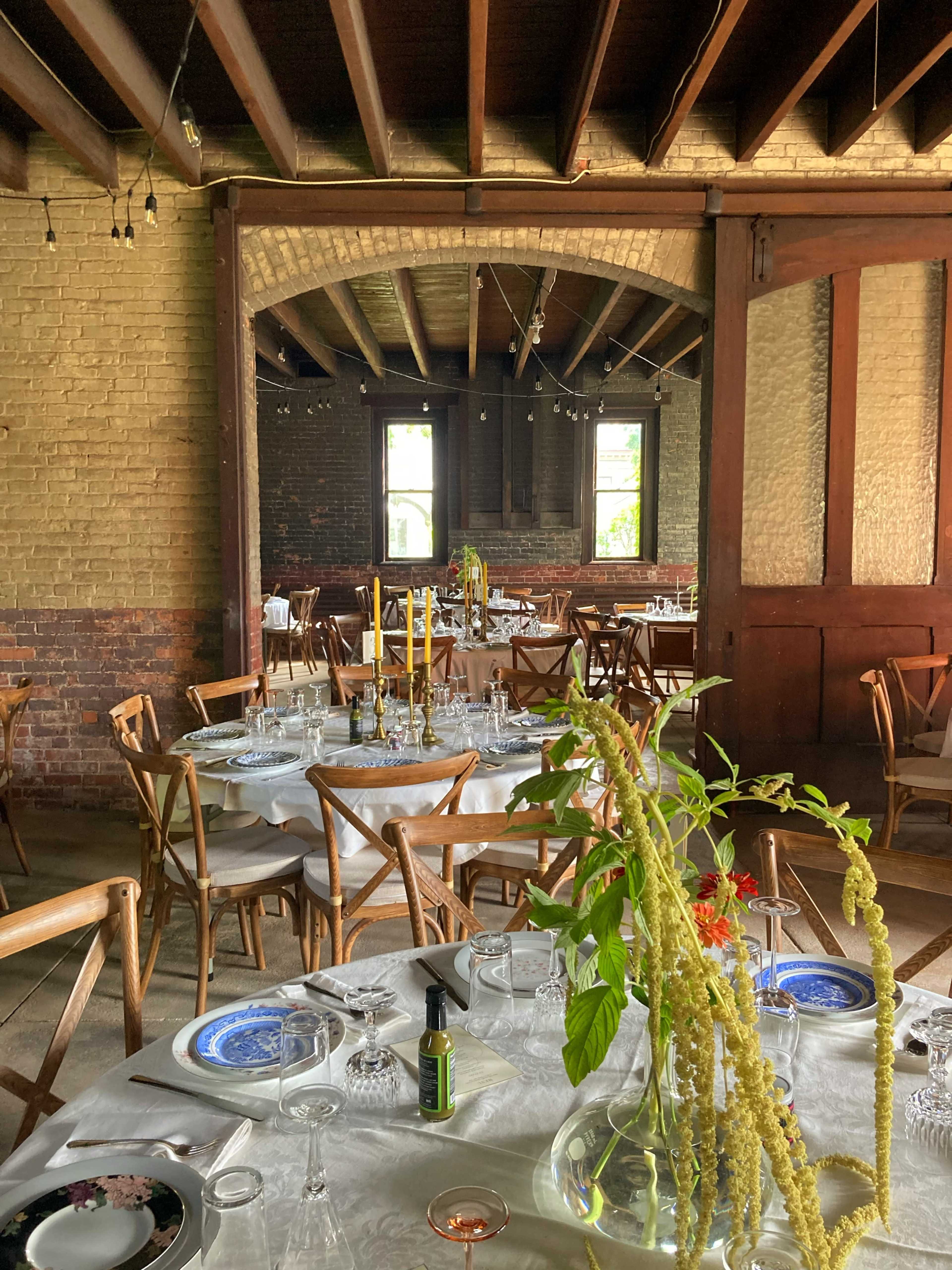 The image shows a dining area with wooden furniture, neatly set tables, and decorative plants in vases, under a ceiling with exposed beams.