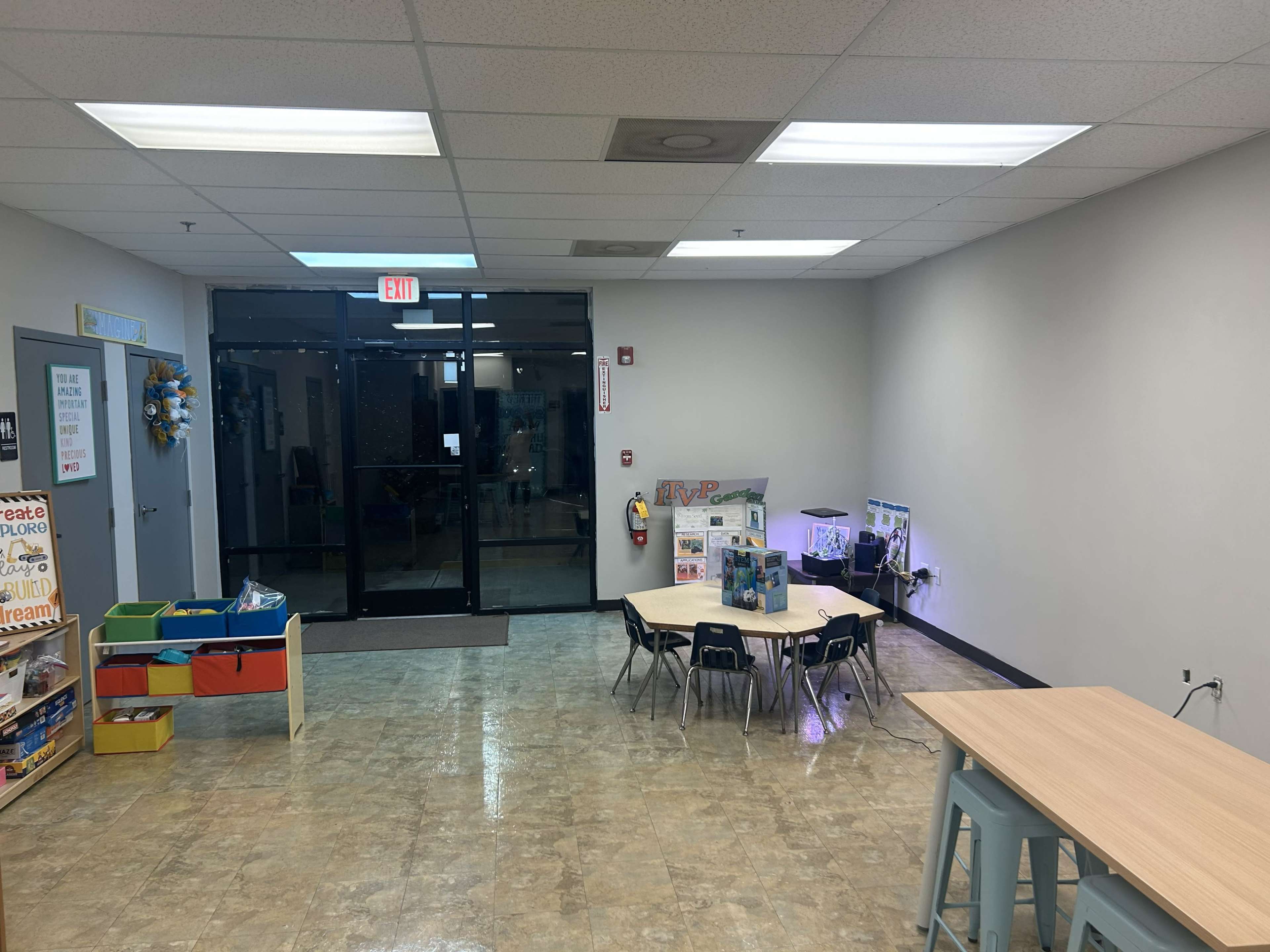 The image shows an empty classroom or play area with tables and chairs, a large entrance door, and some educational displays on the walls.
