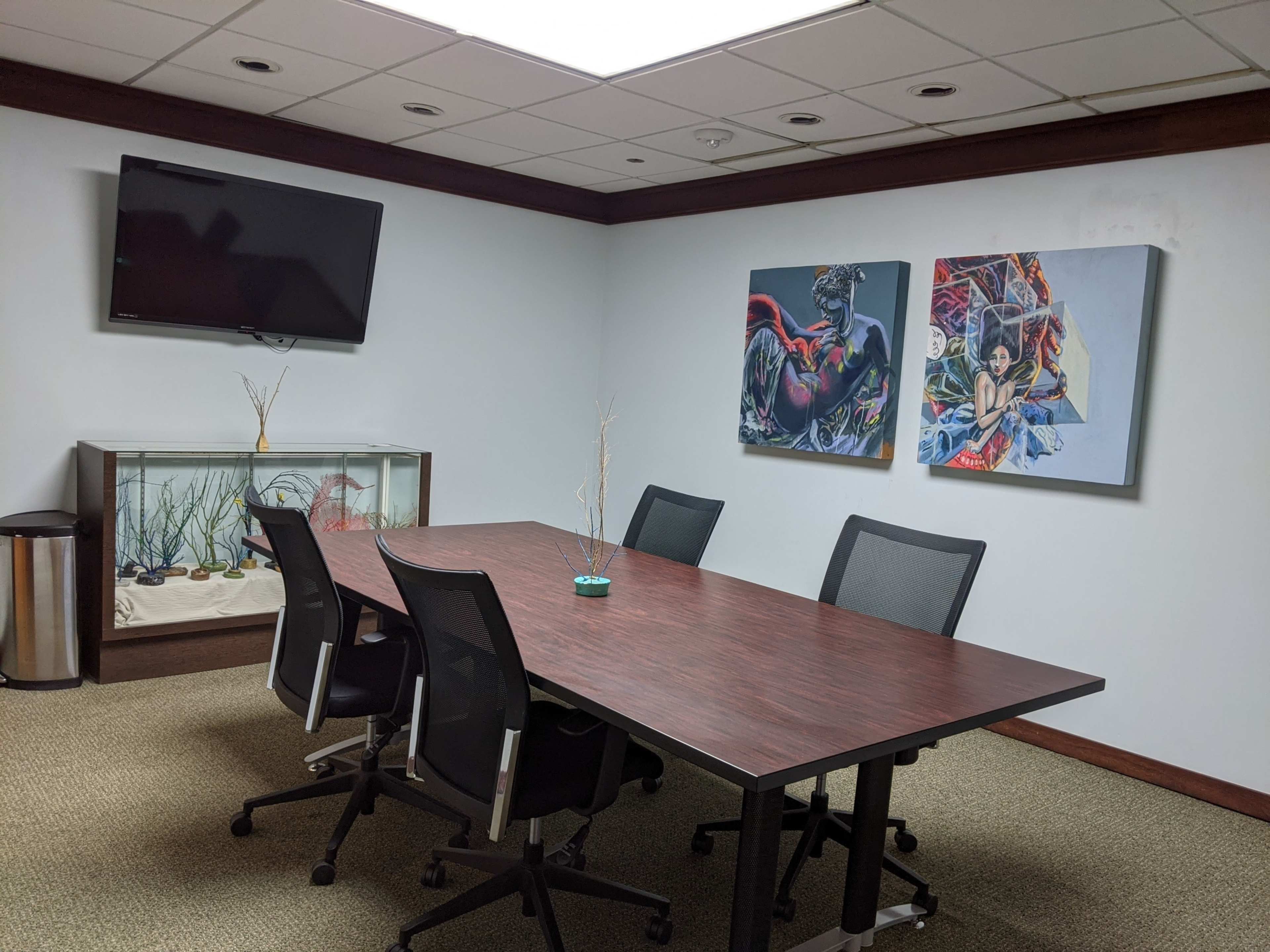 The image shows a conference room with a large wooden table surrounded by black chairs, a television mounted on the wall, and two framed artworks hanging beside a fish tank.